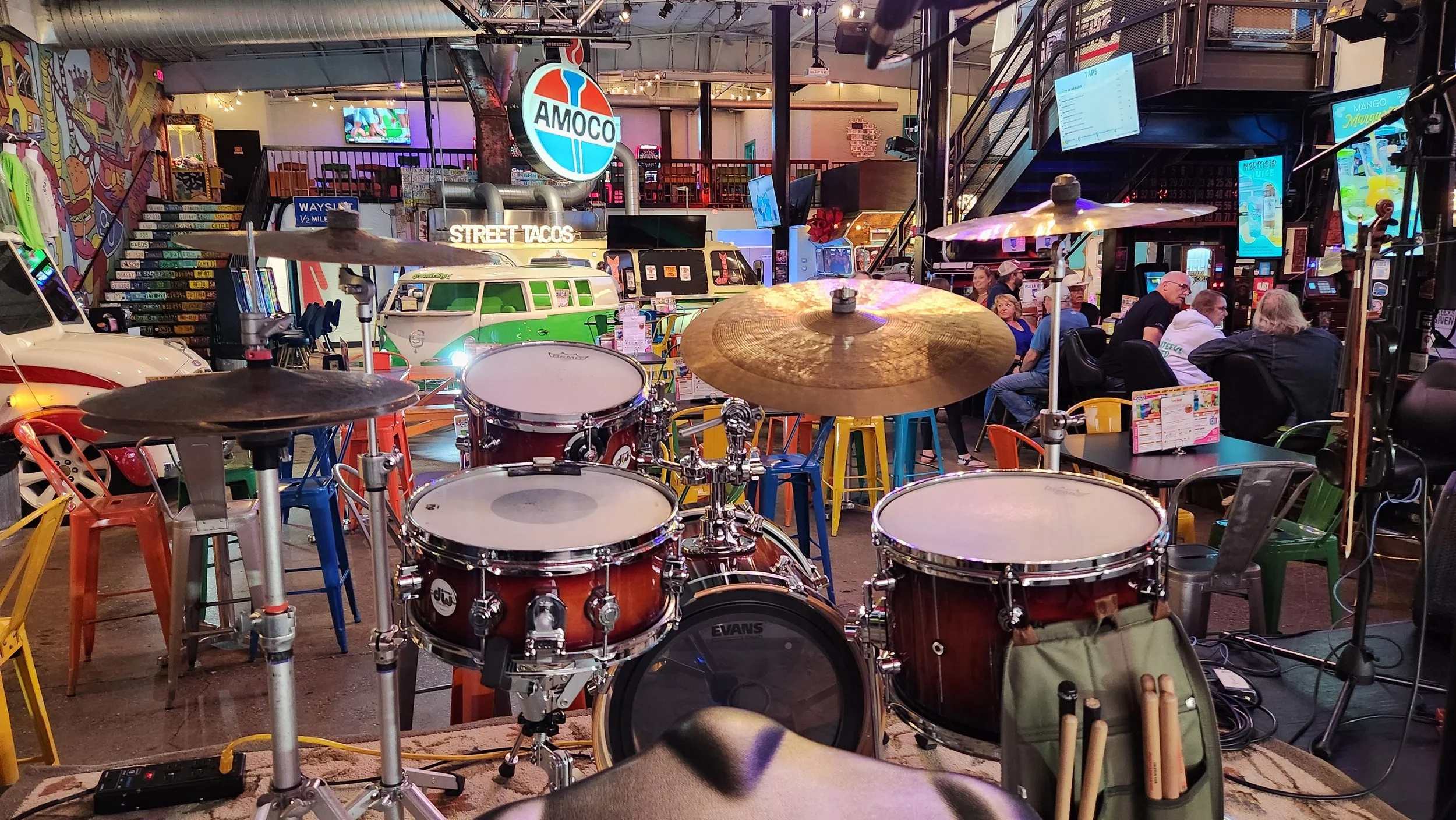 Inside a lively restaurant or bar with a drum set in the foreground, colorful chairs, vintage cars, neon signs, and people socializing at tables.