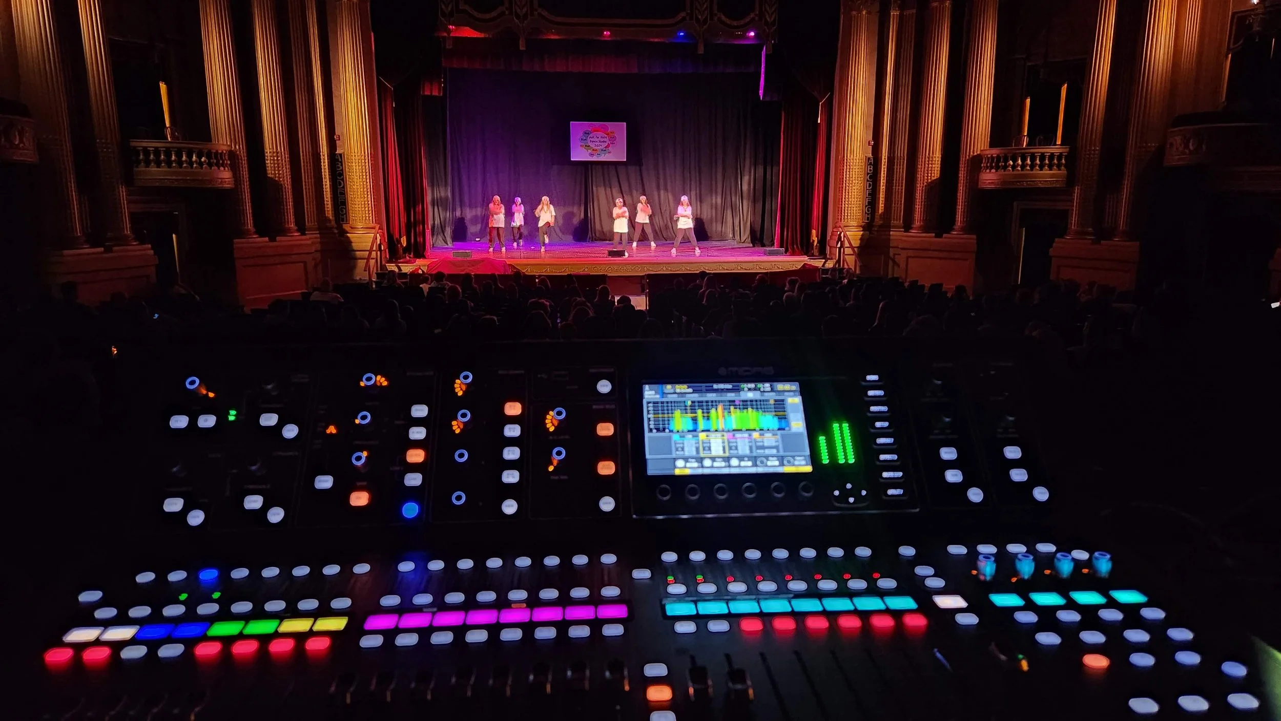 View from a soundboard overlooking a theater stage with performers dancing in front of a black curtain, with an audience in the dark.