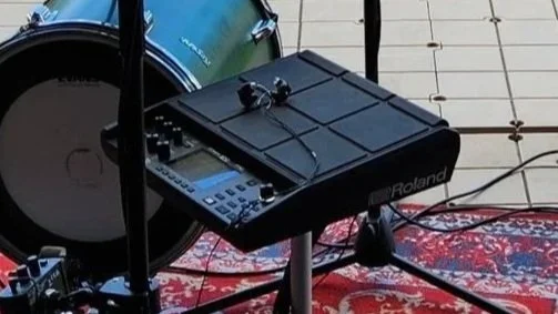 Music production equipment including a Roland drum pad, electronic drum kit, and a pair of headphones on a stand, set up on a patterned rug.