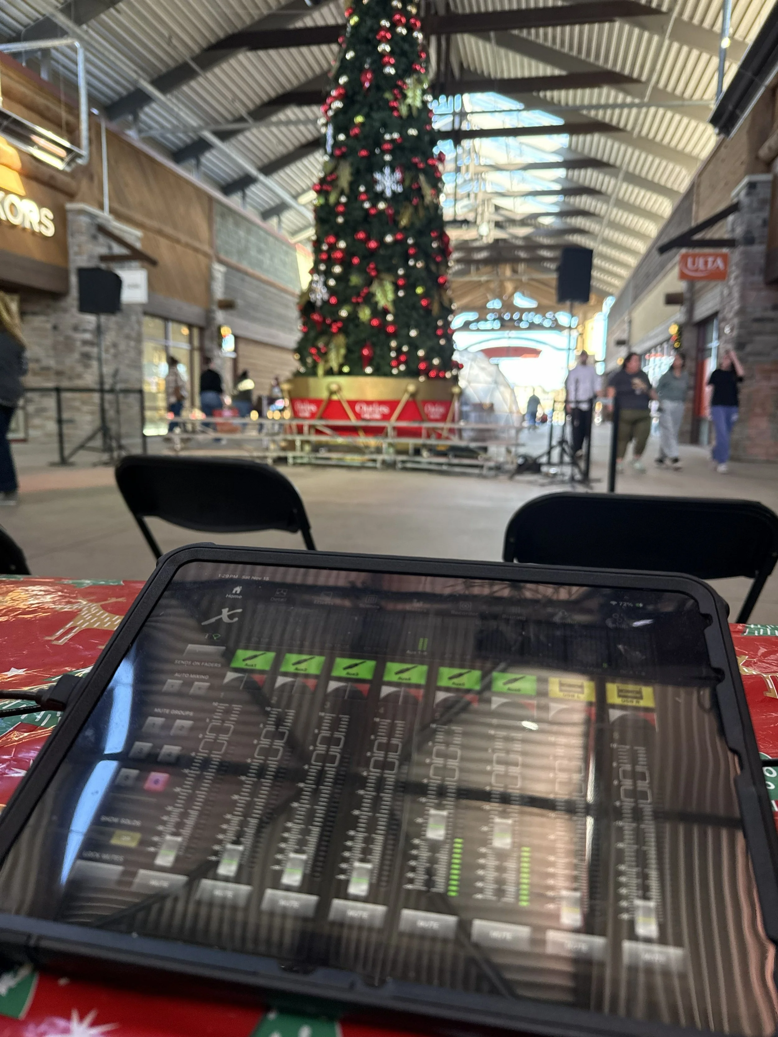 A DJ mixing console with colorful lights and a digital screen, set up on a stage in a dark room with an audience watching a performance.