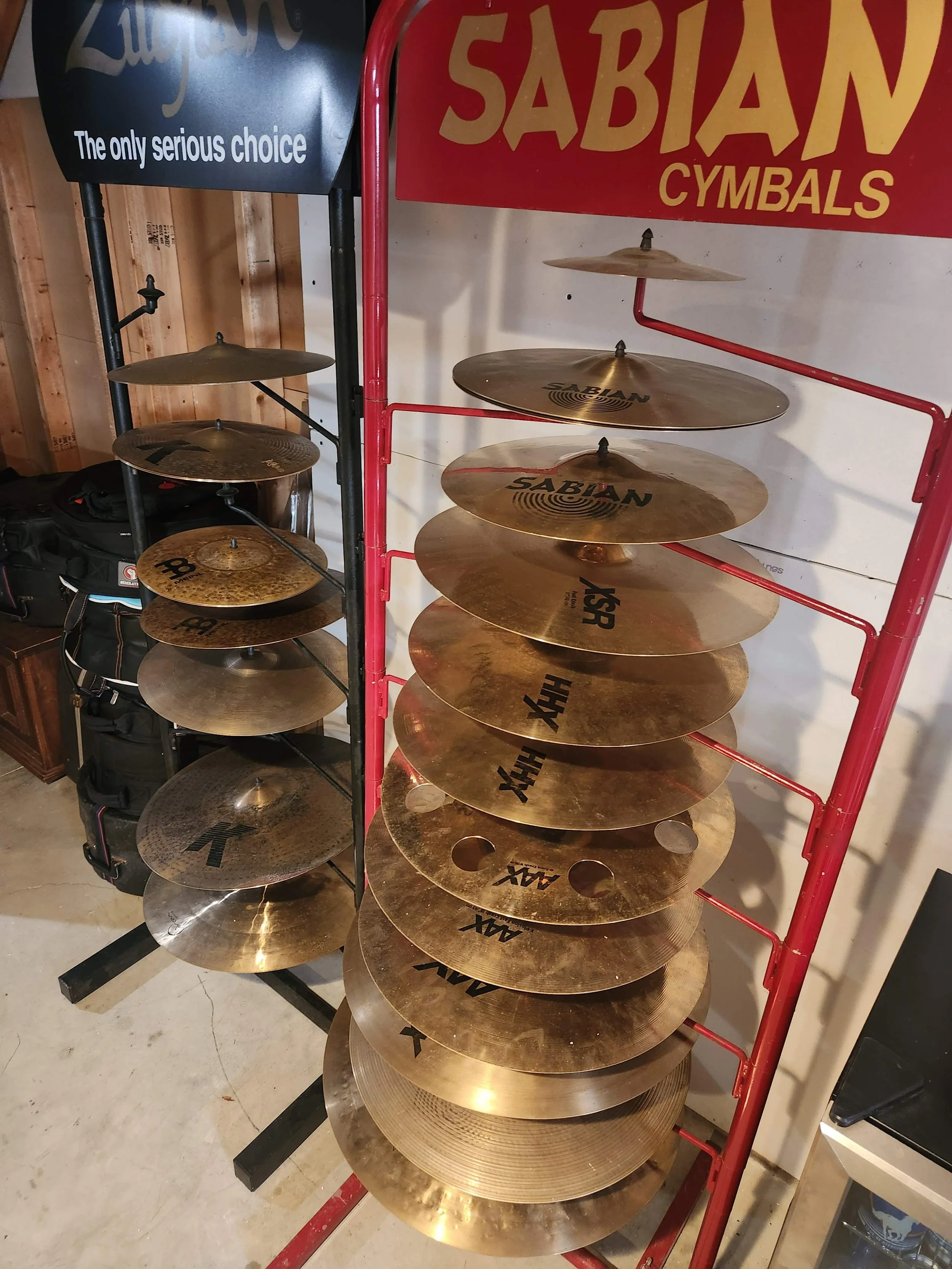 Two cymbal displays, one black stand with Sabian cymbals and another black stand with AAX cymbals, in a music store.