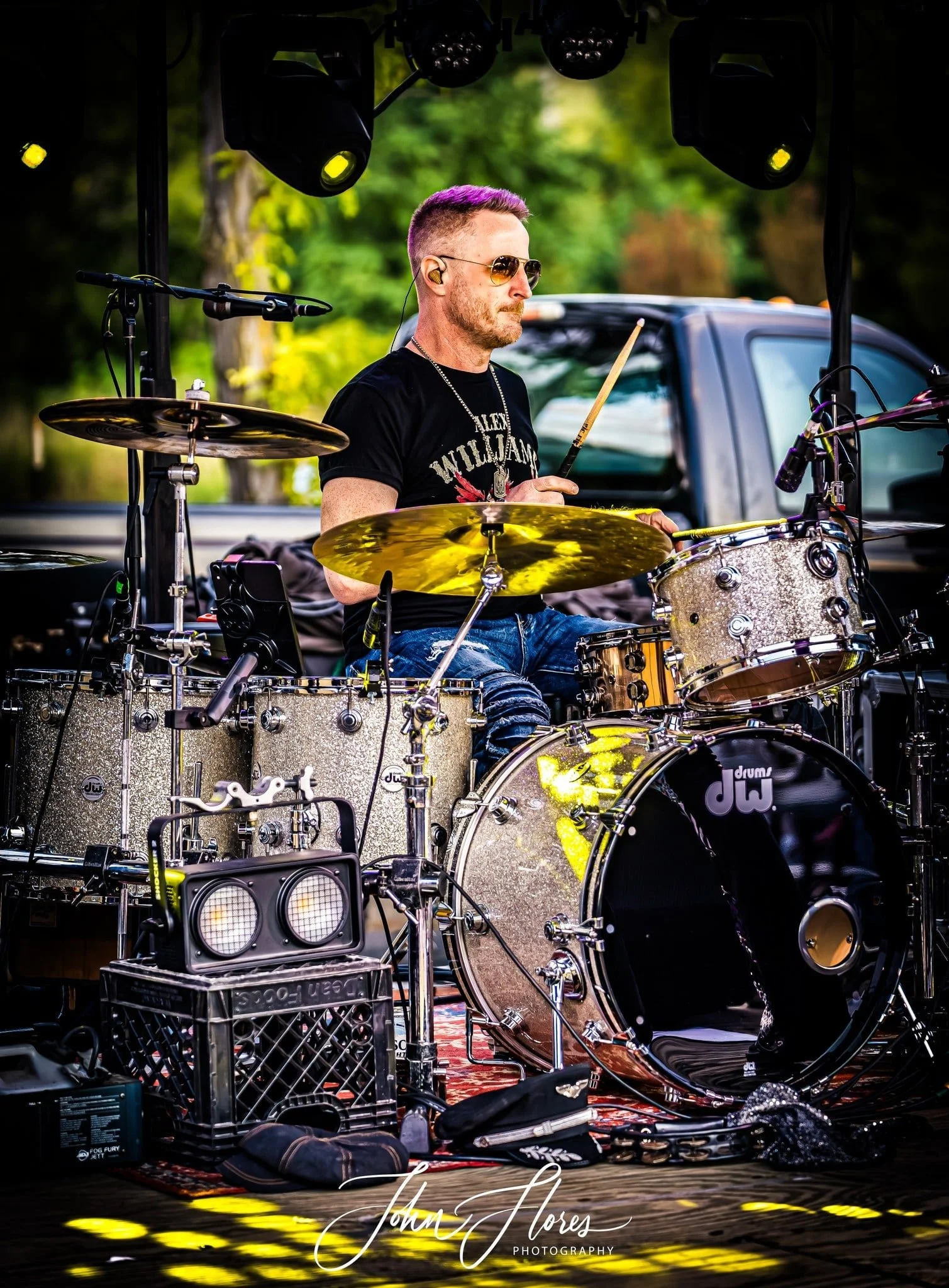 A male drummer playing a drum set during an outdoor performance, wearing sunglasses and a black t-shirt, with trees and a truck in the background.