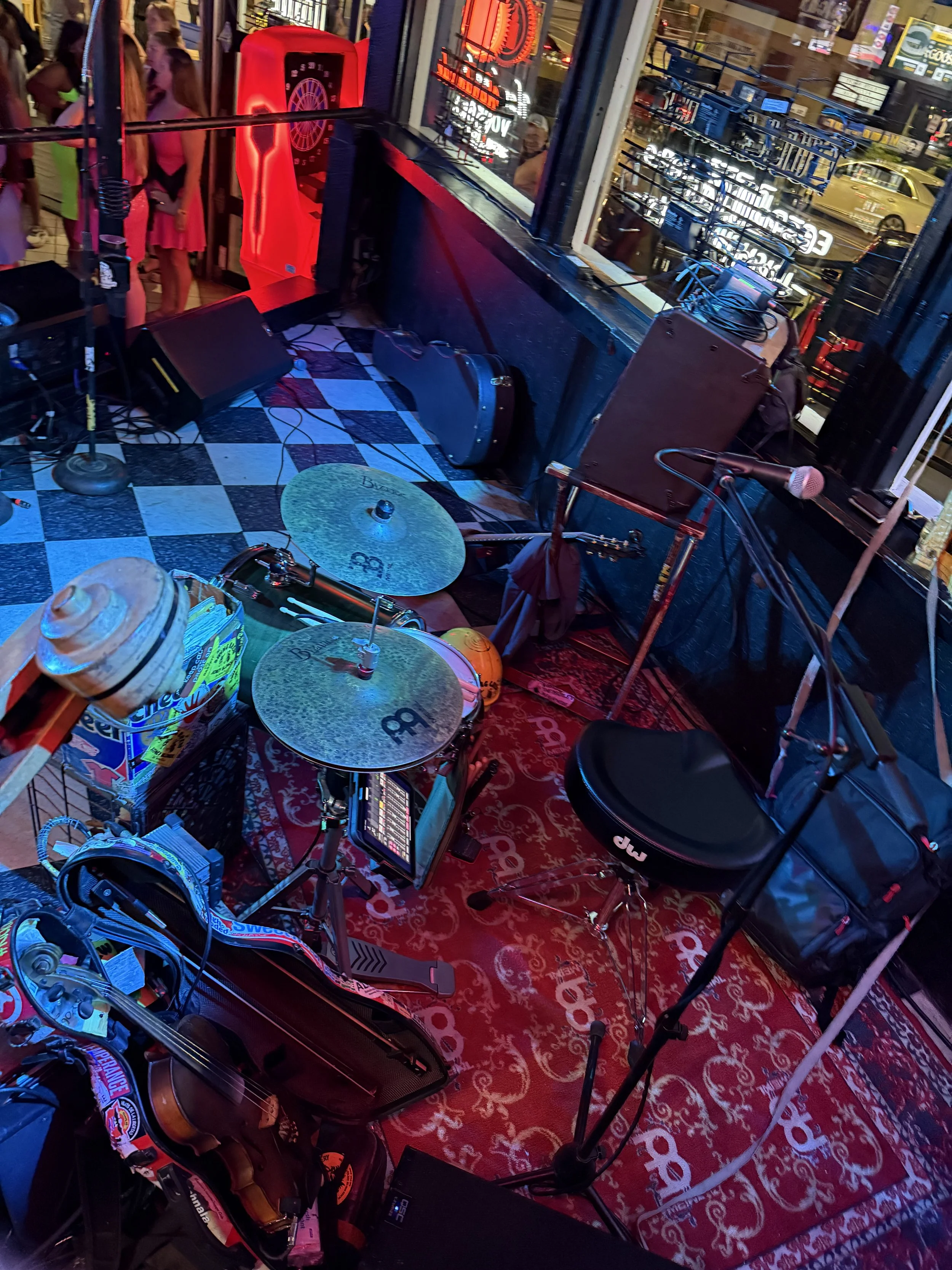 A small stage setup with a drum set, microphone, and other musical equipment in a bar or nightclub, with people visible outside through the window.