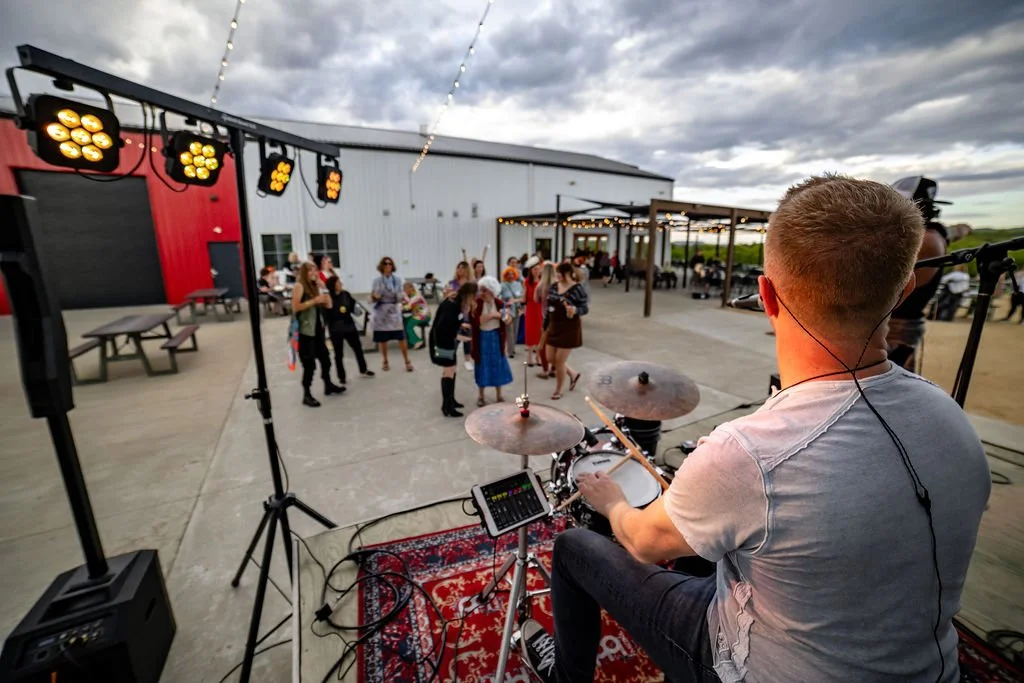 A musician playing drums at an outdoor event with people gathered in the background, under string lights, during evening or late afternoon at a farm or open venue.
