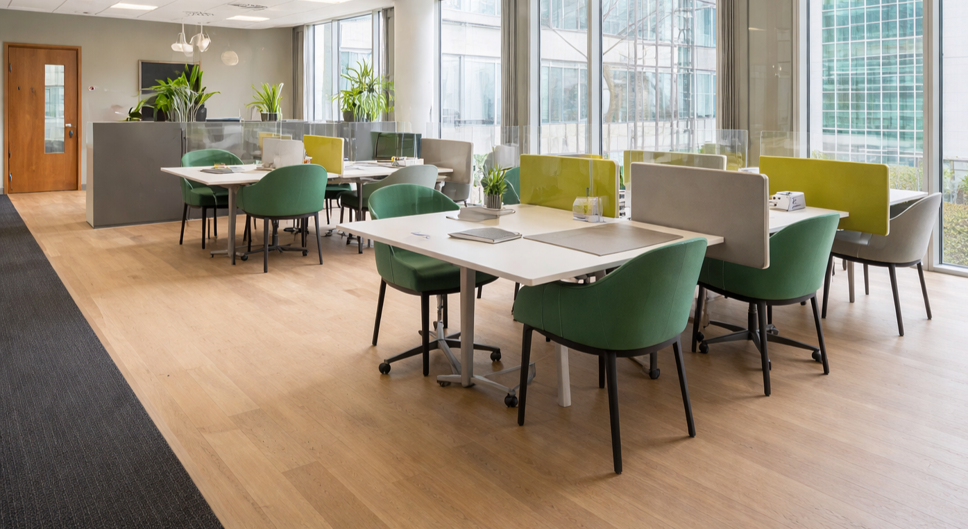 Modern office break room with wooden floors, large windows, green chairs, white tables, and plants.