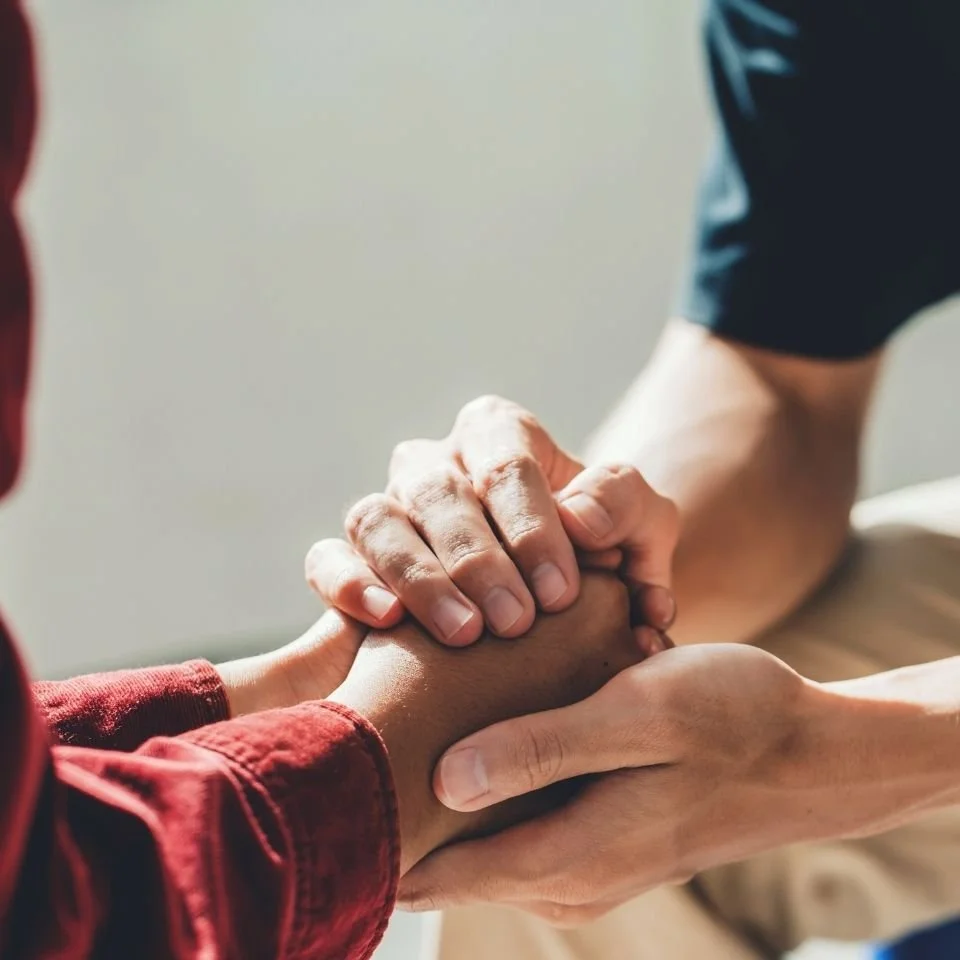 Two people holding hands, one with a darker skin tone and the other with a lighter skin tone, in a comforting or supportive gesture.