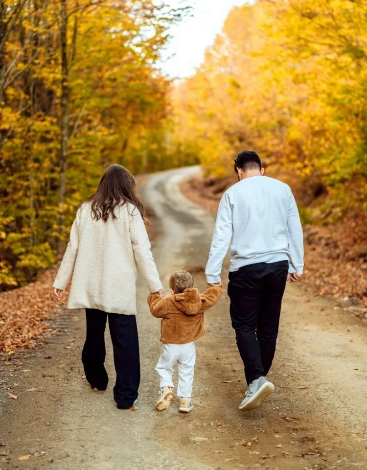 A family of three walking down a dirt road in a fall forest, holding hands, with colorful autumn trees on either side.