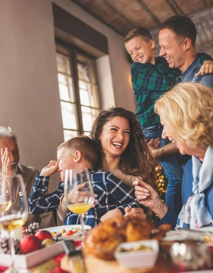 Family gathered around dining table laughing and hugging during a holiday meal.
