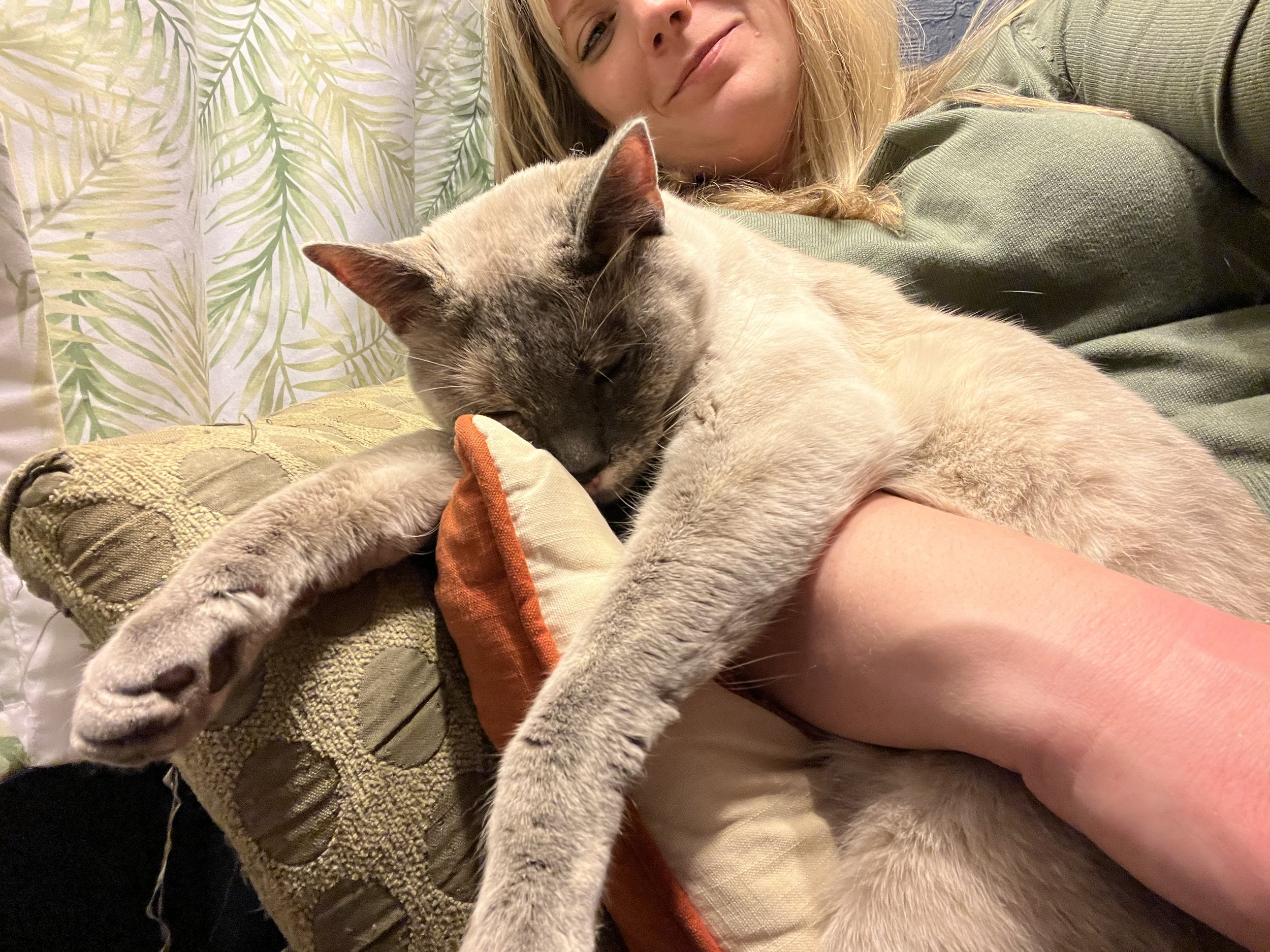 A woman lying on a sofa with a Siamese cat cuddled against her and sleeping.