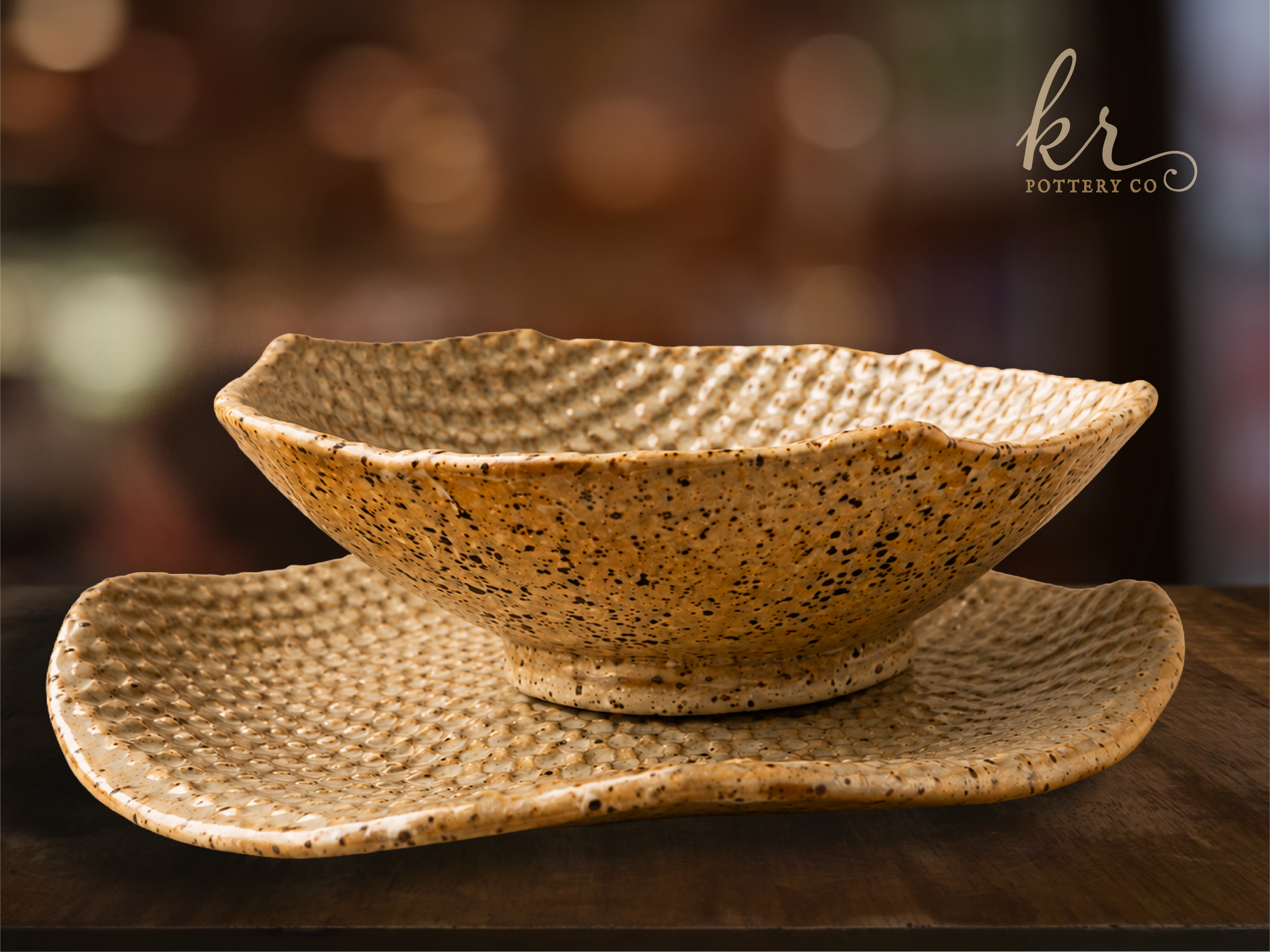 Stacked beige and black speckled ceramic bowls and plates in a display, with a blurred background and a logo that reads 'kr Pottery Co'.