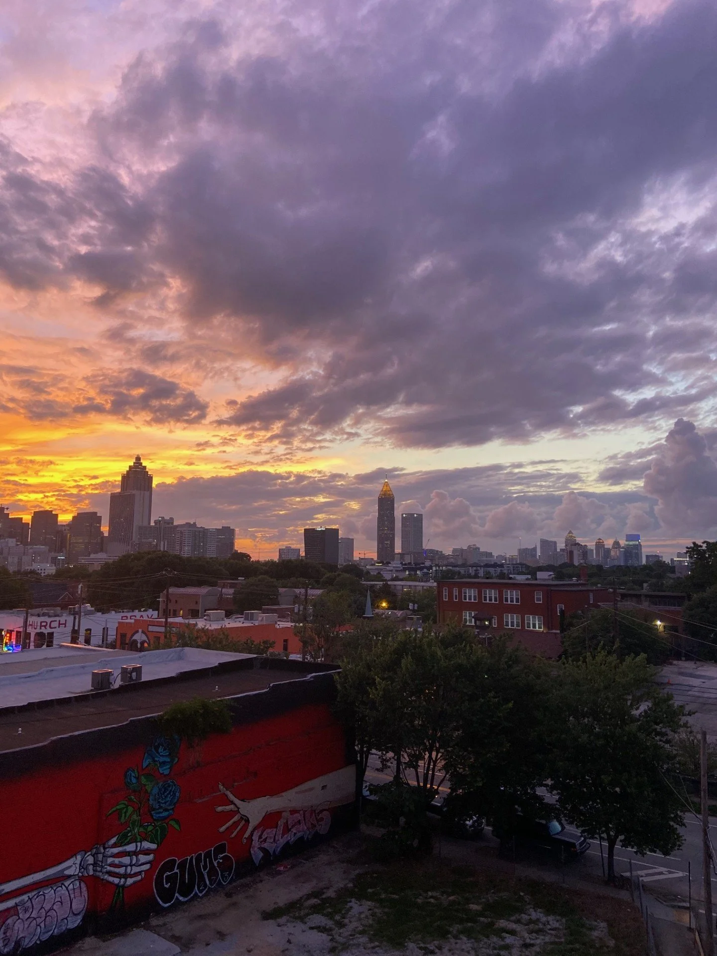 Sunset over Atlanta skyline with purple and orange clouds in the sky. Buildings of various heights, including tall skyscrapers, are visible. In the foreground, a mural of a skeletal hand holding a flower is painted on a red building.