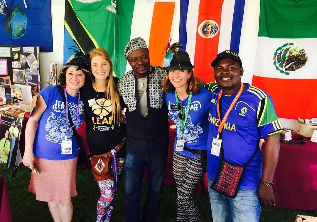 Group of five smiling people wearing African-style clothing and hats, standing in front of flags from different countries including Jamaica, Mexico, and others, at an international event or festival.