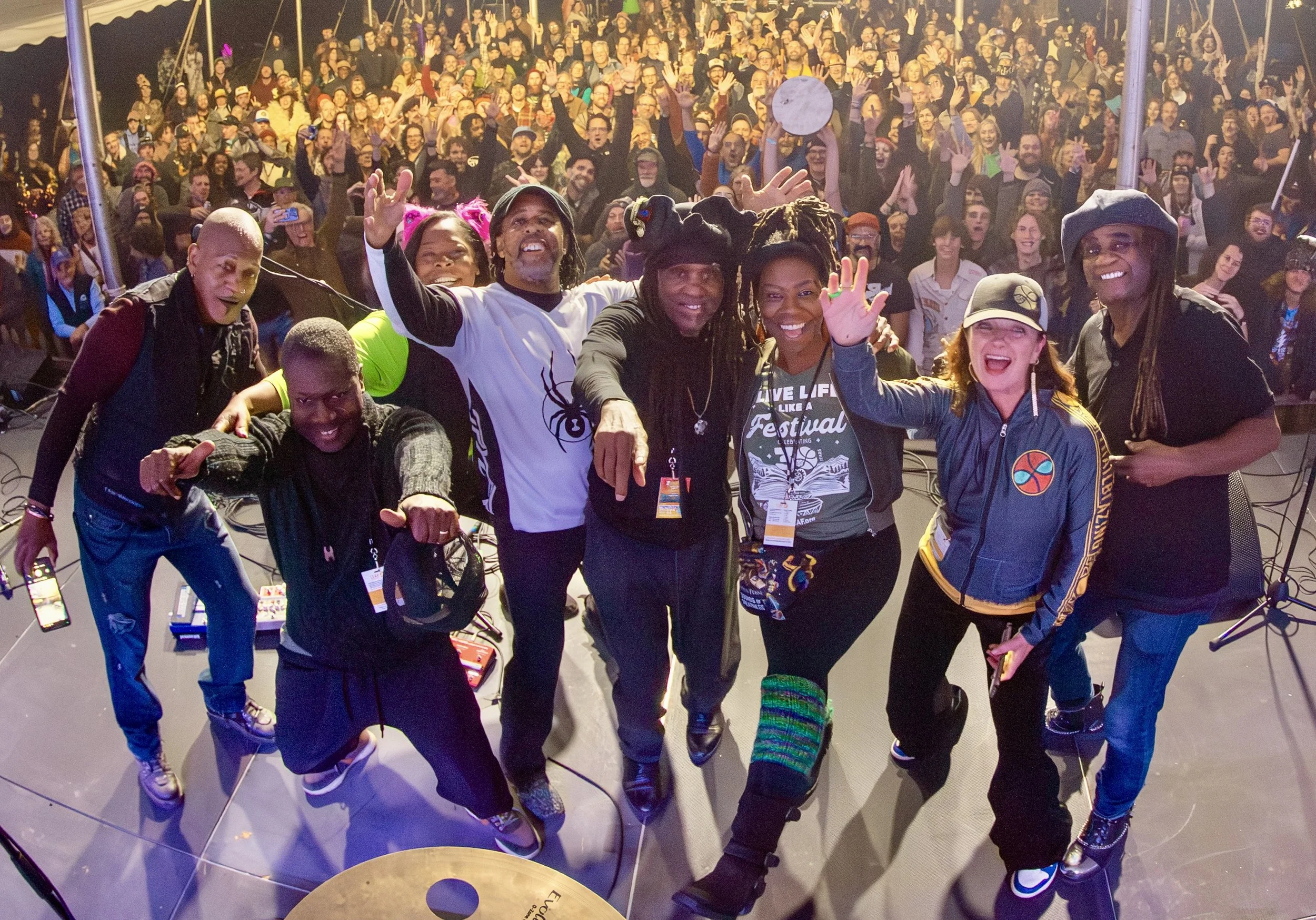 A group of diverse people, including men and women, posing for a photo on a stage with a large crowd in the background at a lively event or concert.