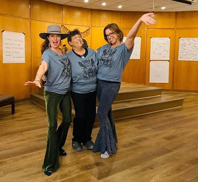 Three women standing together in a room with wooden walls and a small stage, smiling and posing playfully for the camera.