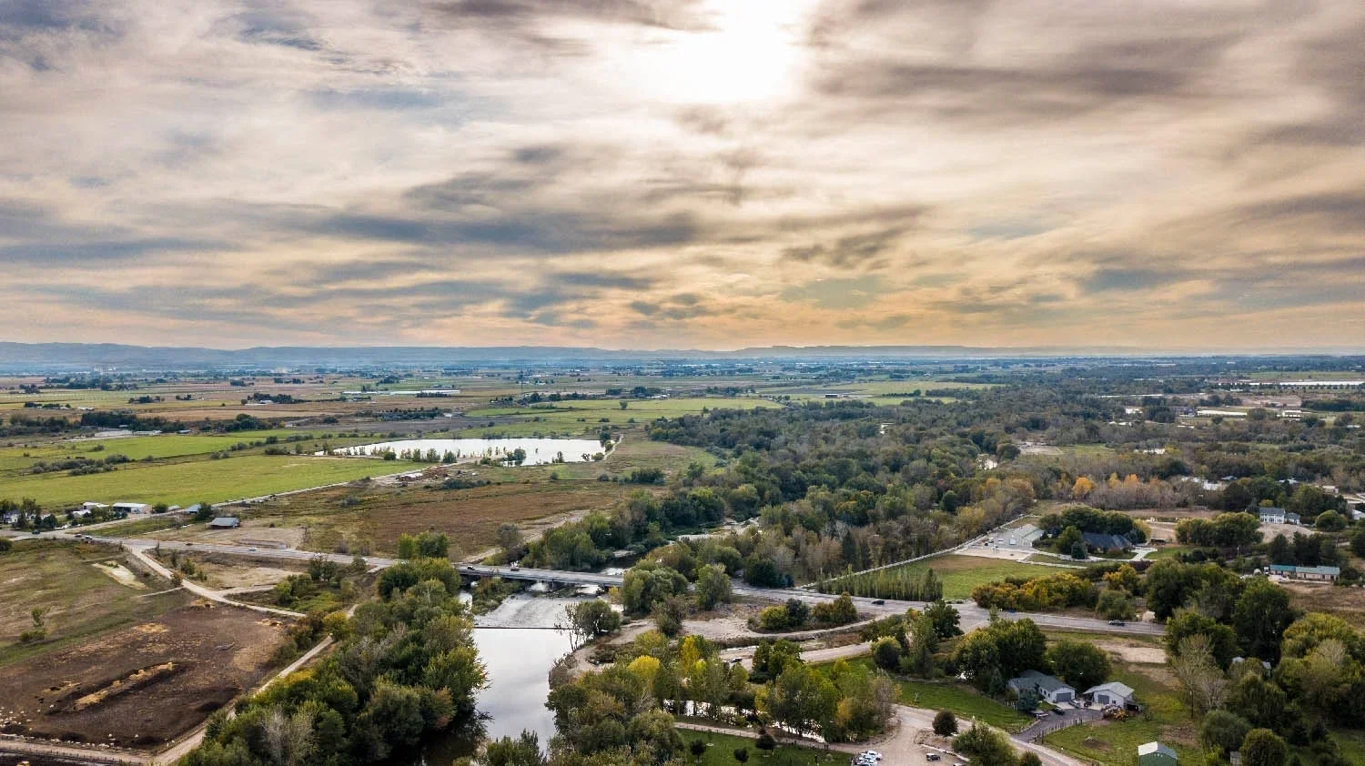 Aerial view of a rural landscape with a river, trees, fields, roads, and a few buildings under a partly cloudy sky at sunset.