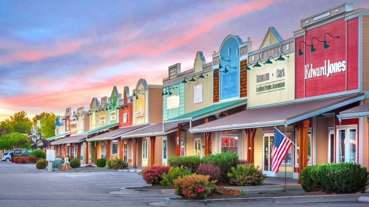 Colorful storefronts in a shopping center at sunset, with an American flag in the foreground and a partly cloudy sky.
