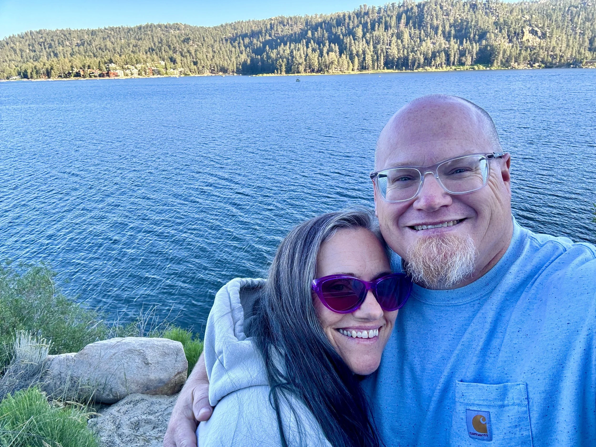 A happy couple takes a selfie by a large blue lake with mountains and trees in the background, on a sunny day.