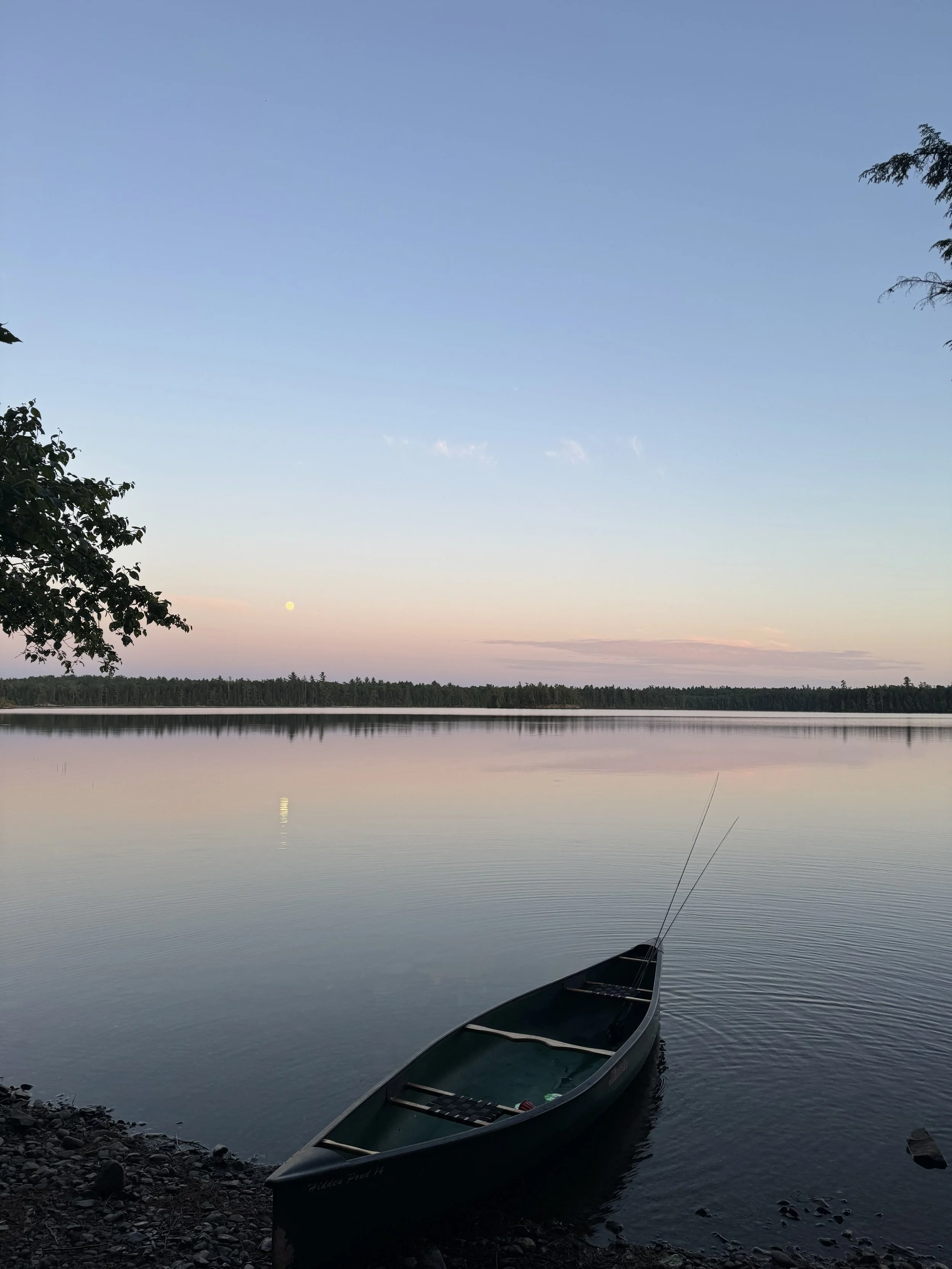 A canoe resting on the rocky shore of a calm lake during sunset or twilight, with trees on either side and a clear sky reflecting on the water.