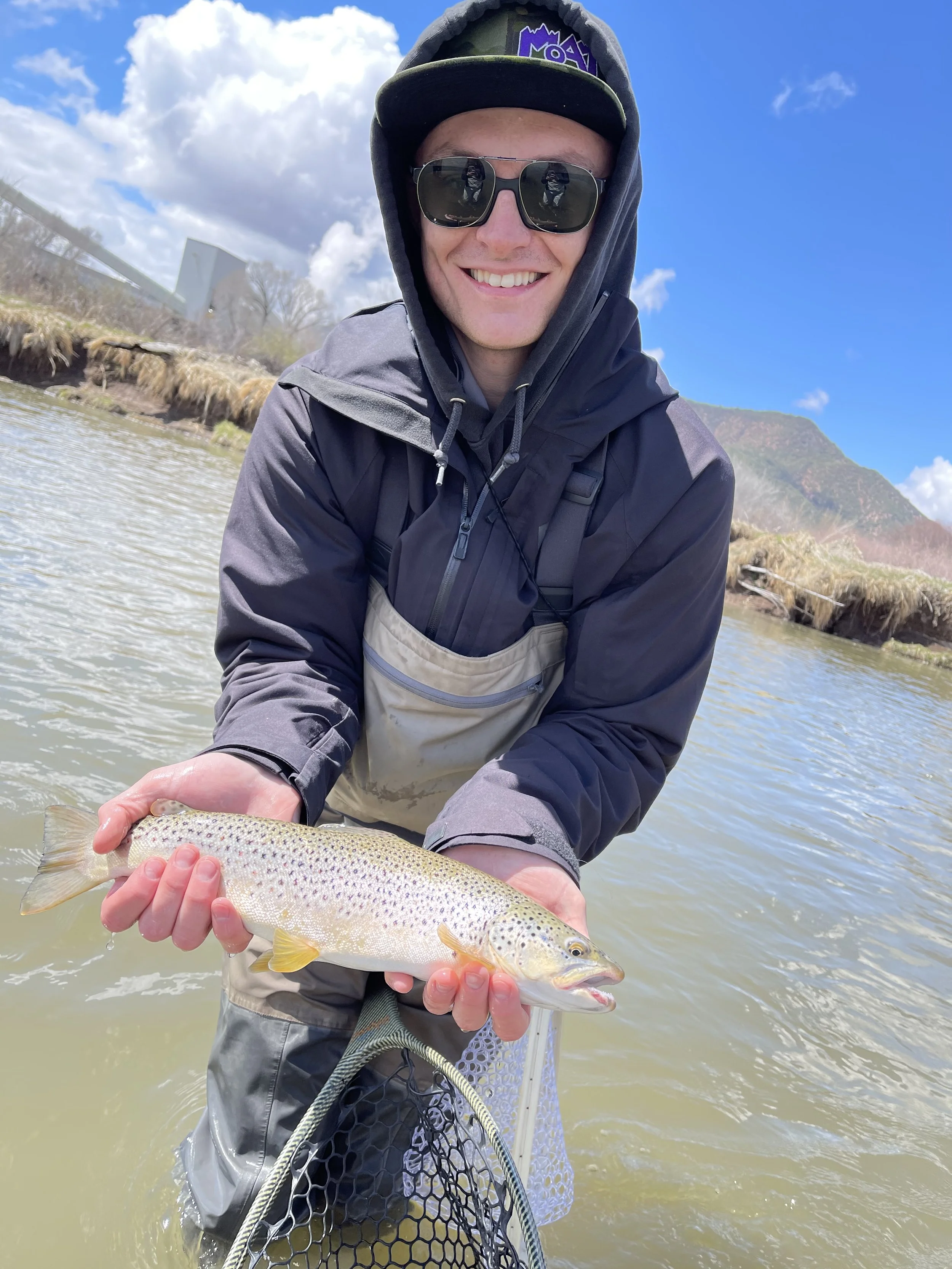 A smiling man in outdoor fishing gear holding a rainbow trout by the river with mountains and a blue sky in the background.