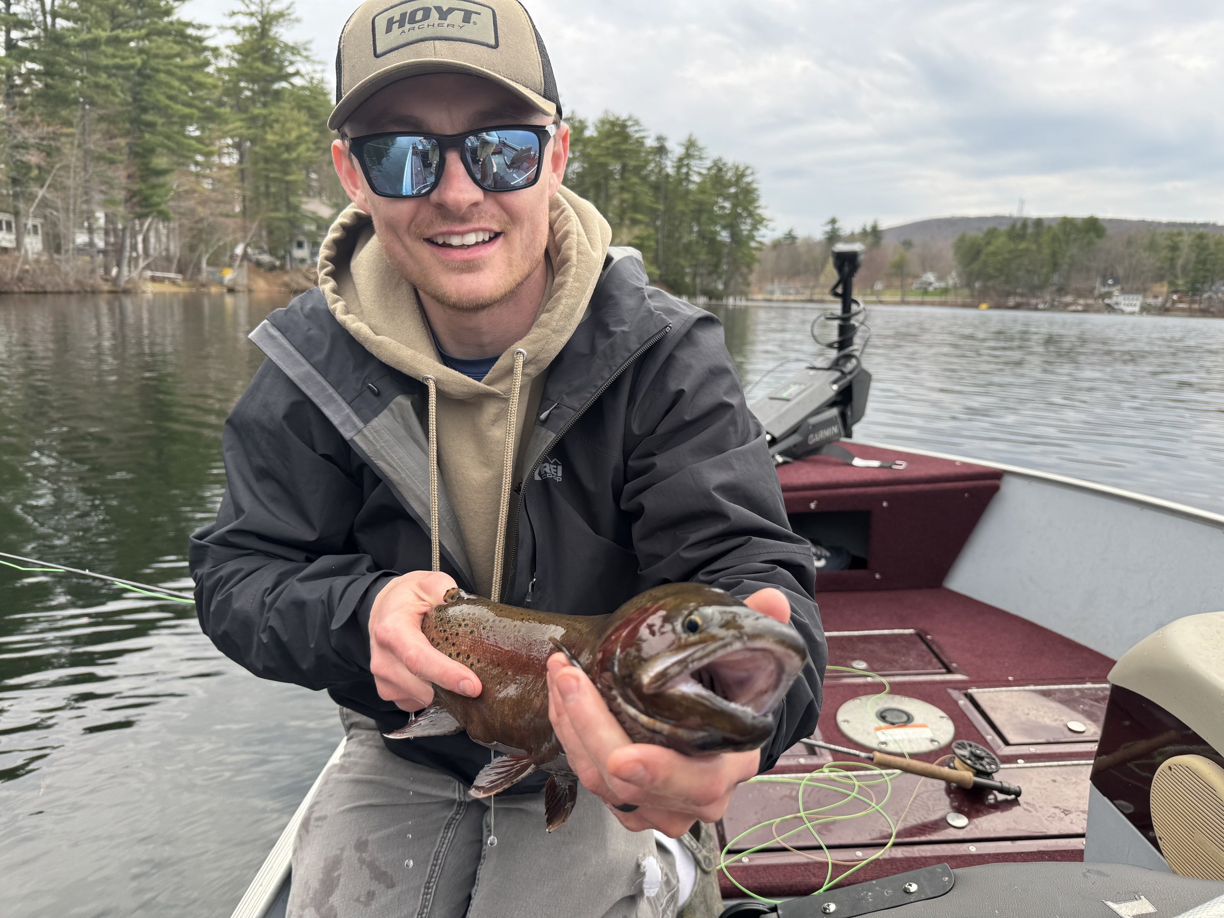 A man smiling while holding a large fish on a boat on a lake, with trees and houses in the background.