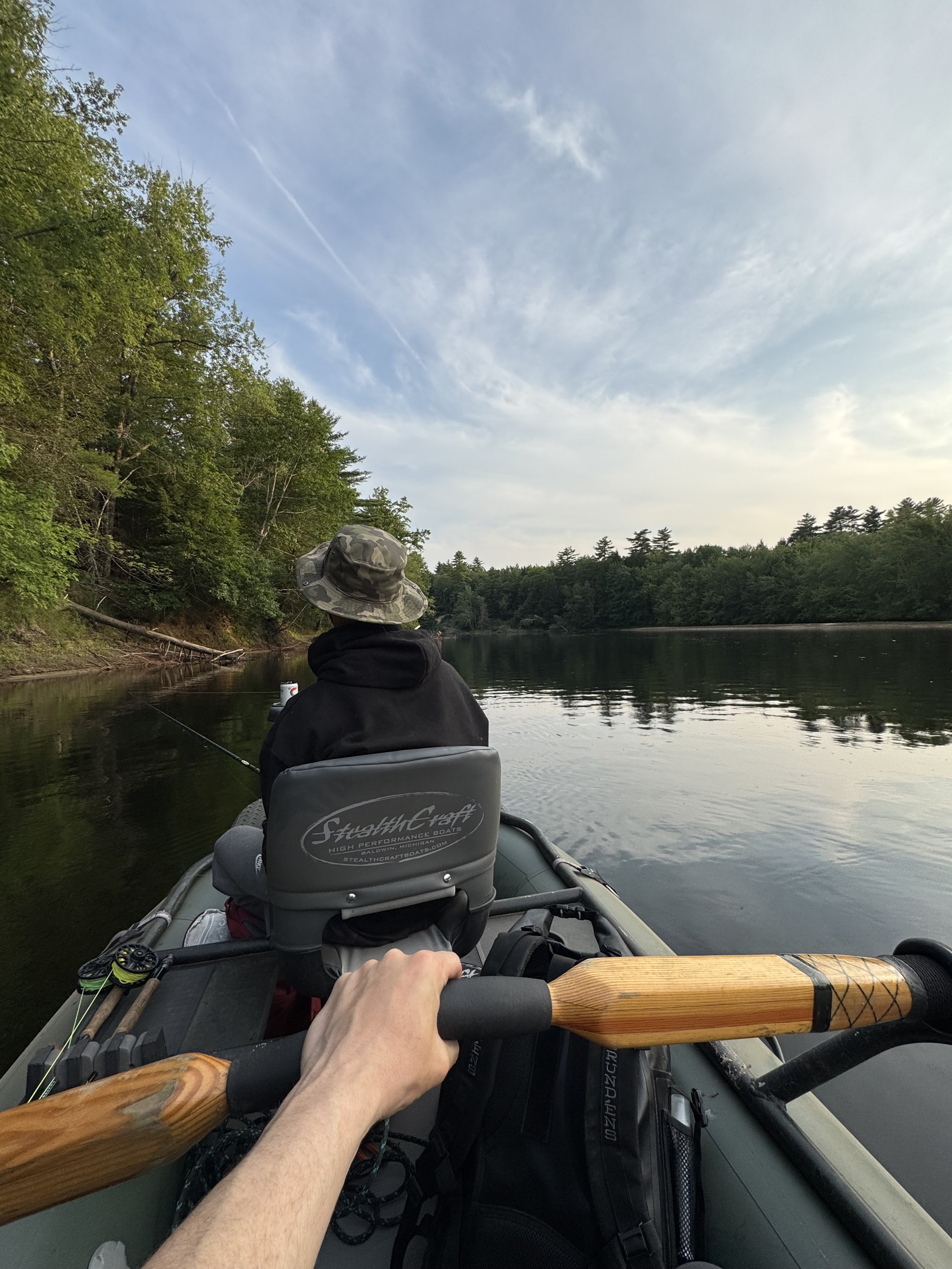 Person paddling an inflatable boat on a river with trees on the banks, fishing, and a partly cloudy sky.