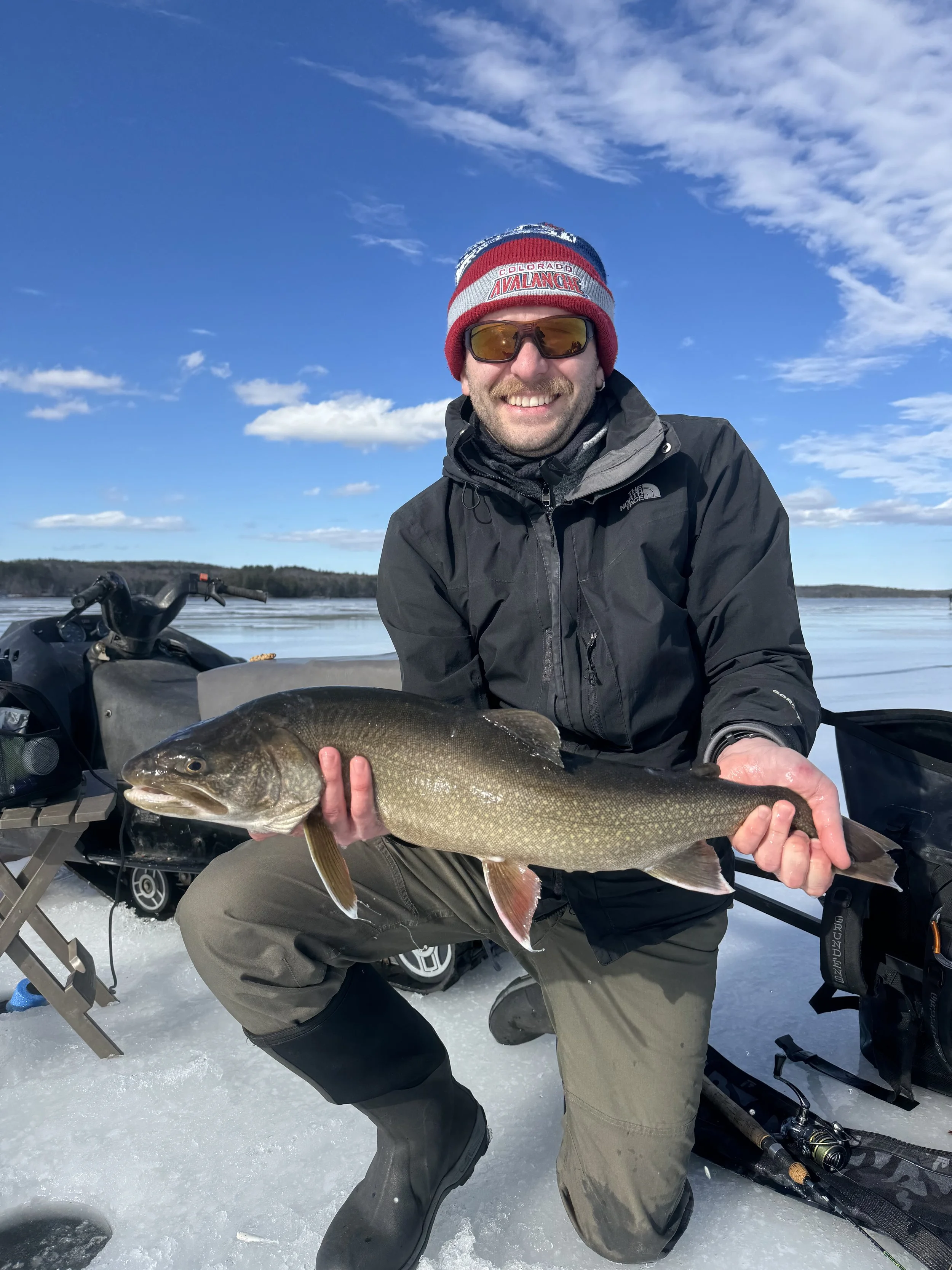 A man in winter clothing and sunglasses kneeling on snow, holding a large fish with both hands, with ice fishing gear and a lake in the background.