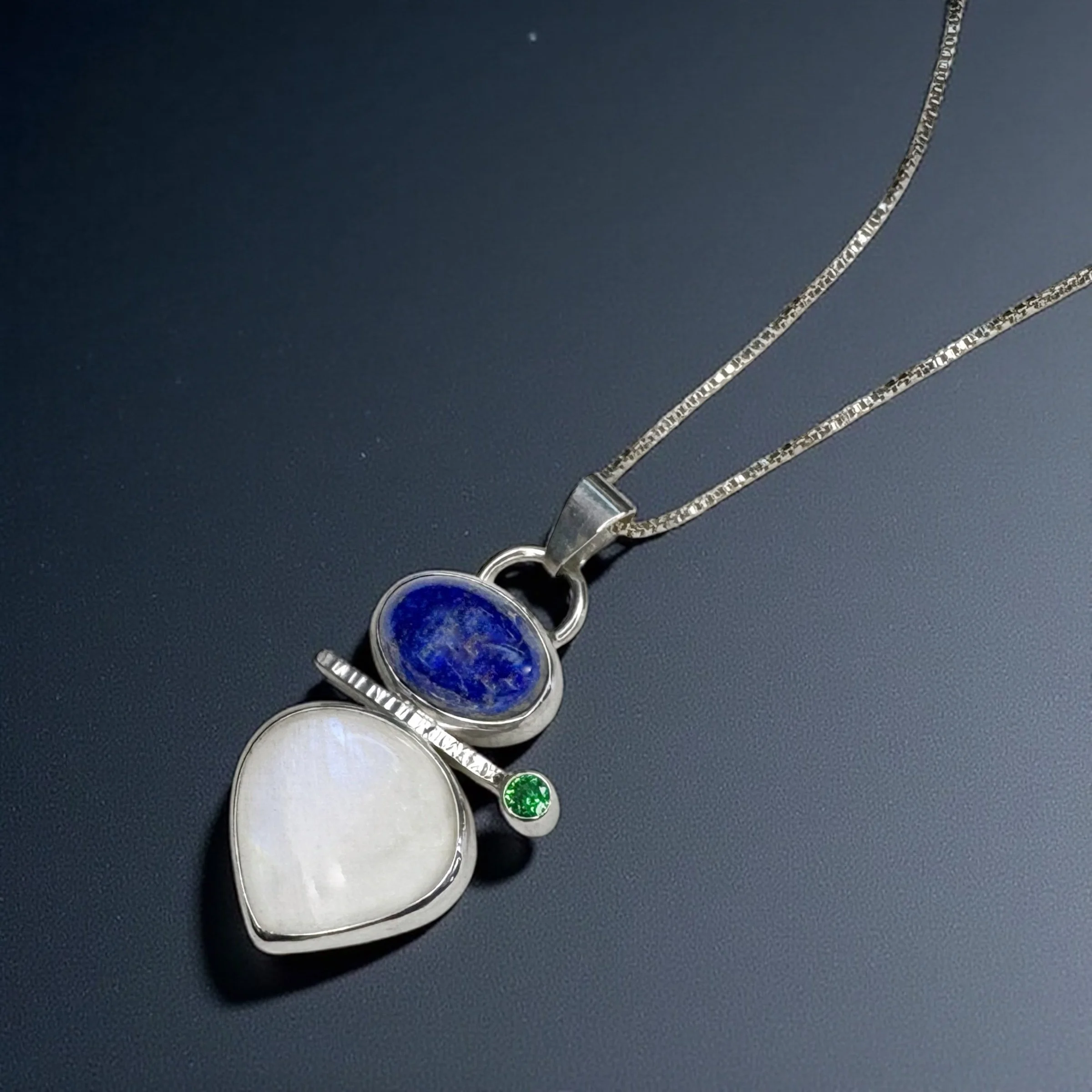 Lapis lazuli and moonstone in a sterling silver pendant with a green cubic zirconia accent.