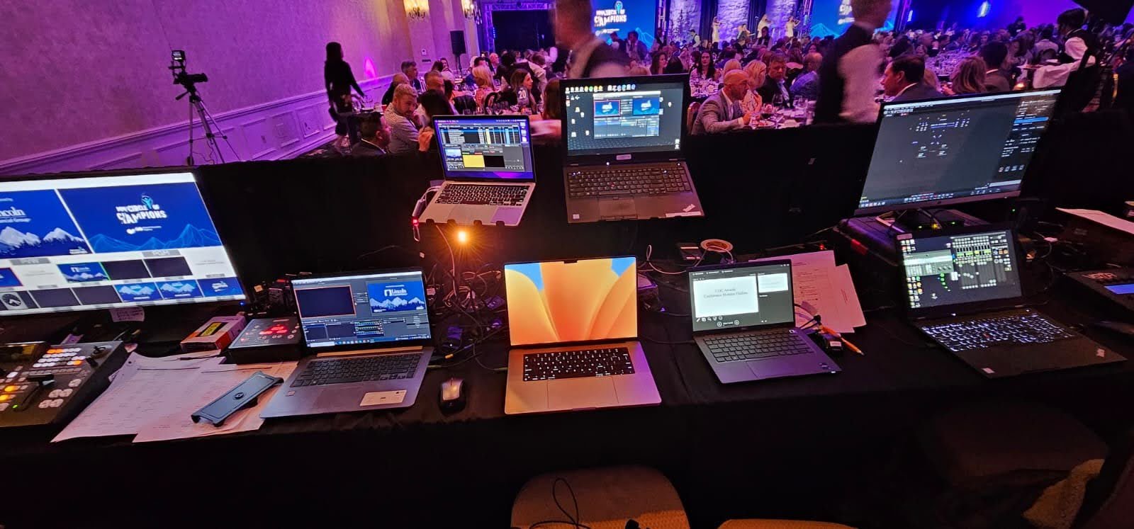 A setup of multiple laptops, monitors, and audio equipment on a table at a conference or event with a large audience in the background.
