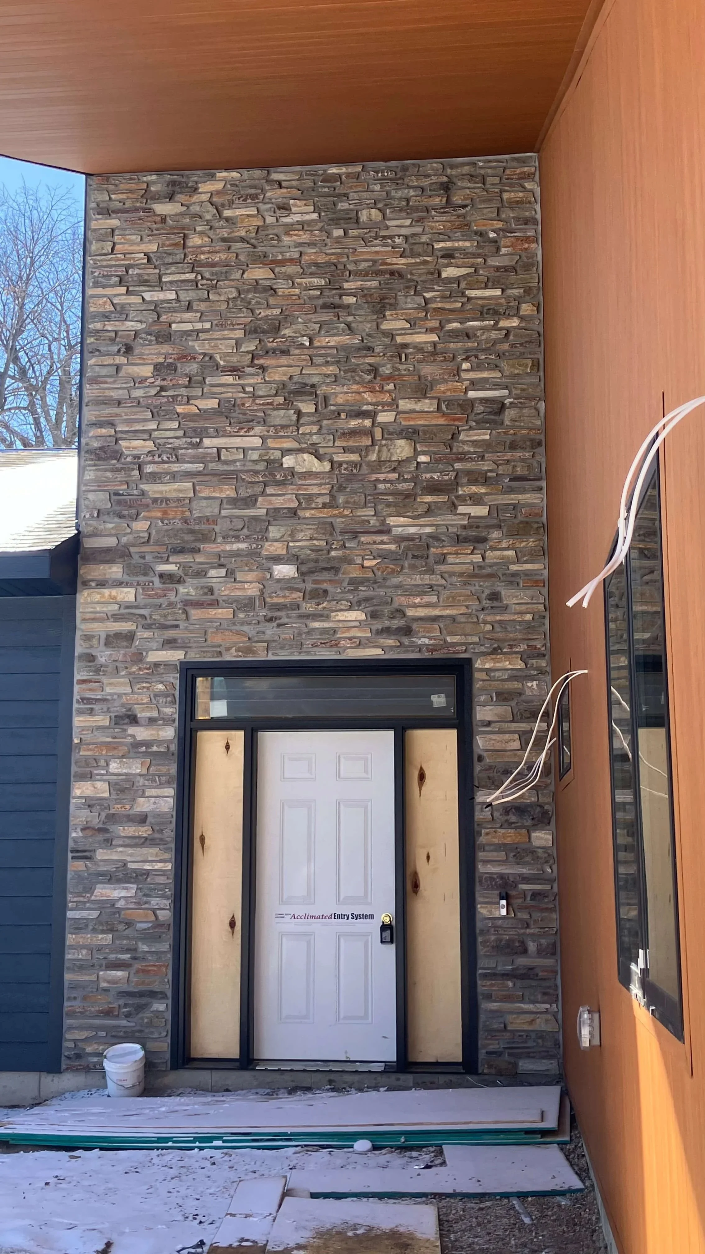 Front door of a house under construction with a stone wall, vinyl siding, and electrical wires hanging on the side.