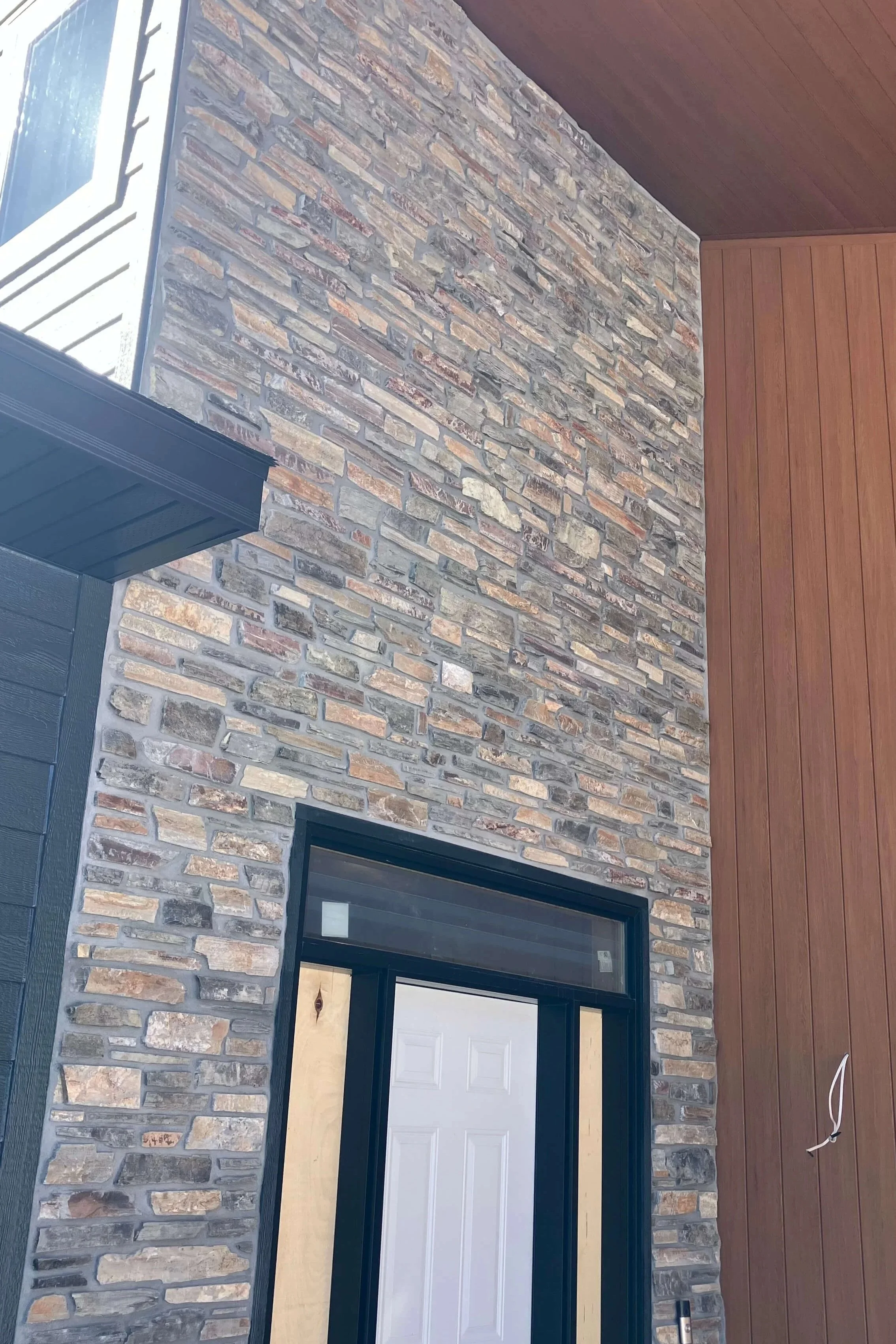 Close-up of a modern building's exterior with a stone wall, wooden paneling, a black-framed window, and a porch roof.