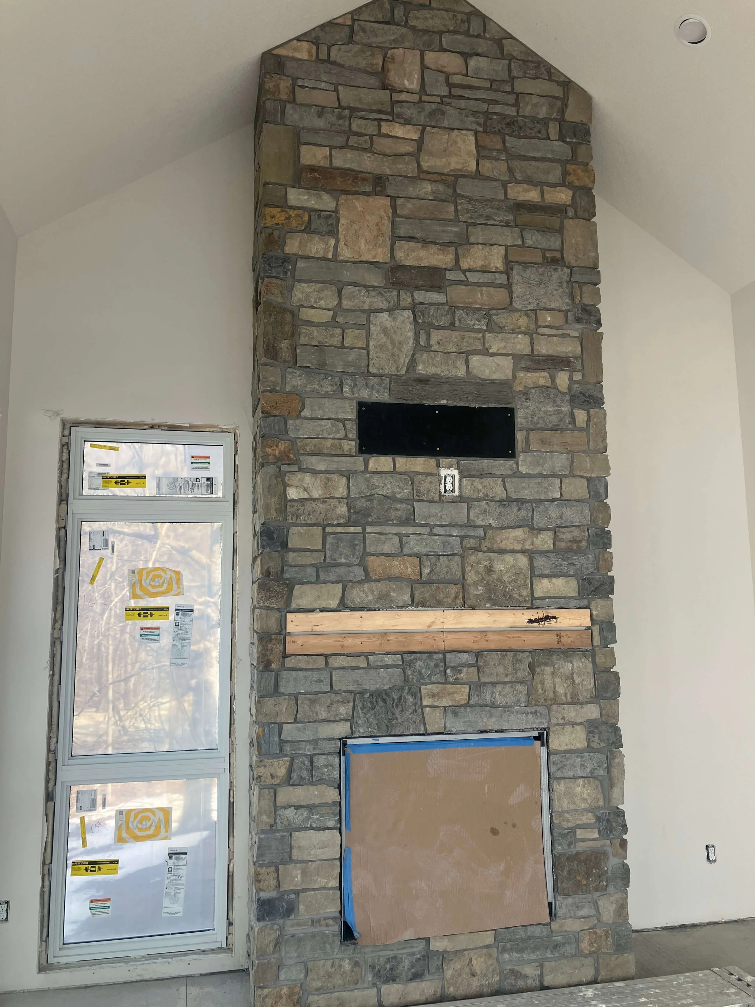 A stone fireplace in an unfinished room, with a window on the left and electrical outlets on the wall. The fireplace has a partially completed hearth and a chimney structure made of large, irregularly shaped stones.