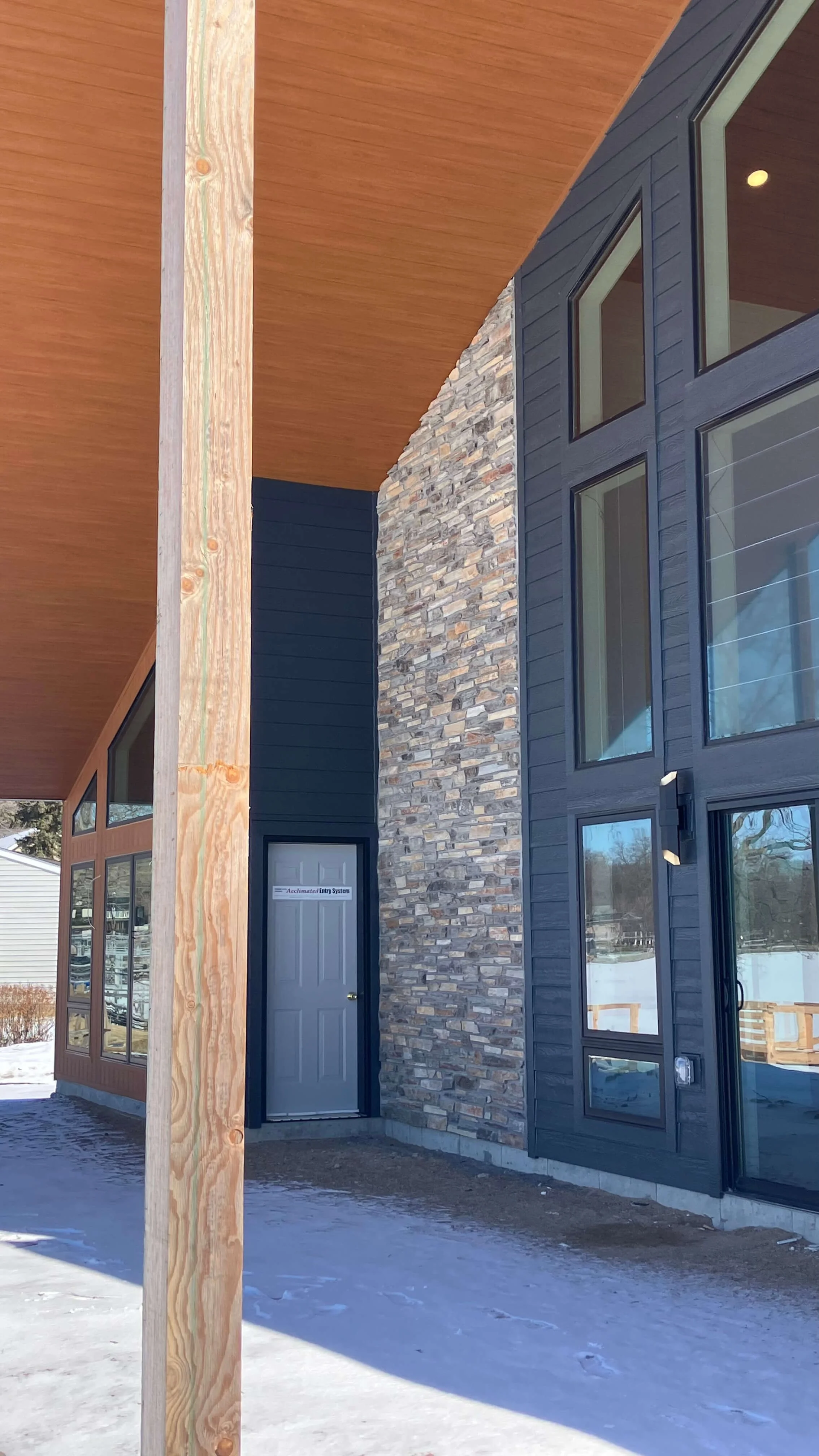 Part of a modern building exterior with a stone wall, large windows, and a door, under a wooden overhang, with snow on the ground.
