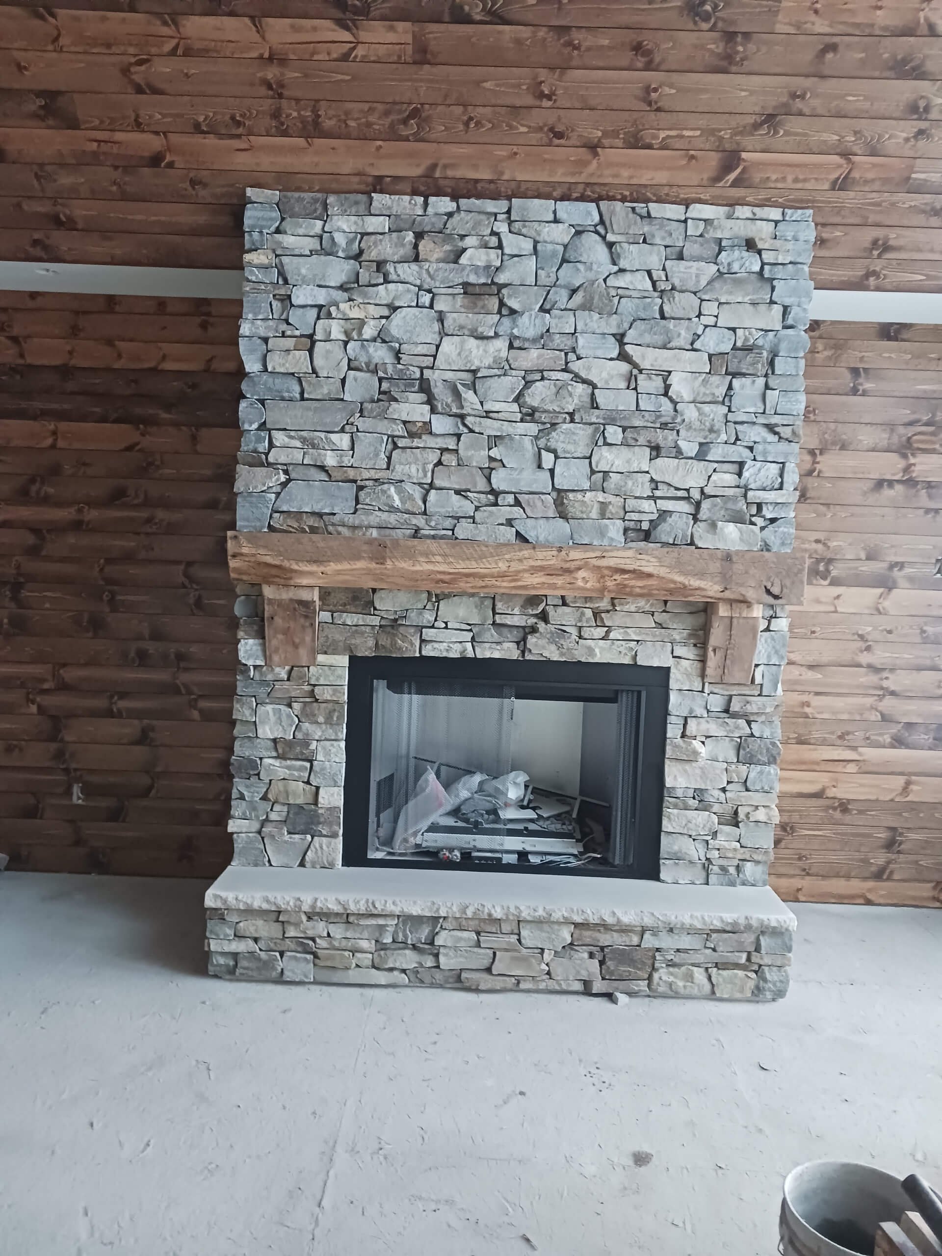 Stone fireplace with wood mantel in a room with wooden ceiling and unfinished concrete floor.