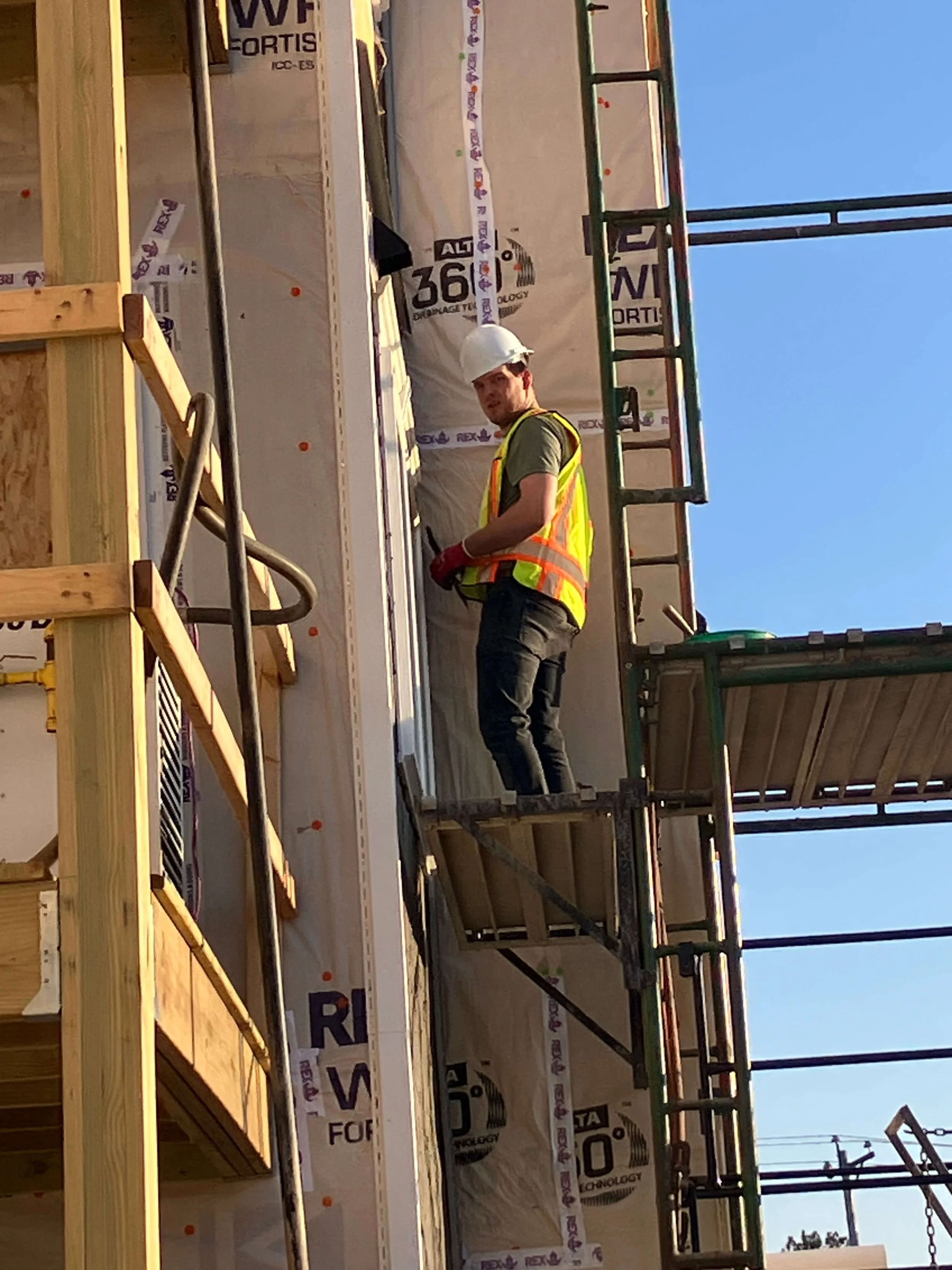 A construction worker wearing a white safety helmet, yellow reflective vest, and dark pants, stands on scaffolding at a building site. The worker is looking towards the camera and appears to be working or inspecting something on the building's exteri
