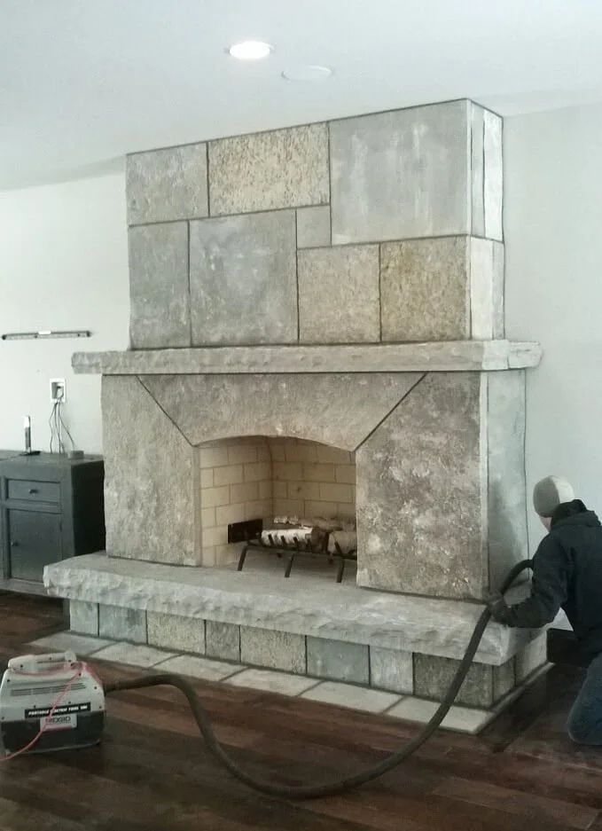 A person is cleaning a large stone fireplace with a vacuum cleaner. The fireplace is made of light-colored stone and has a brick interior, situated on a carpeted floor.