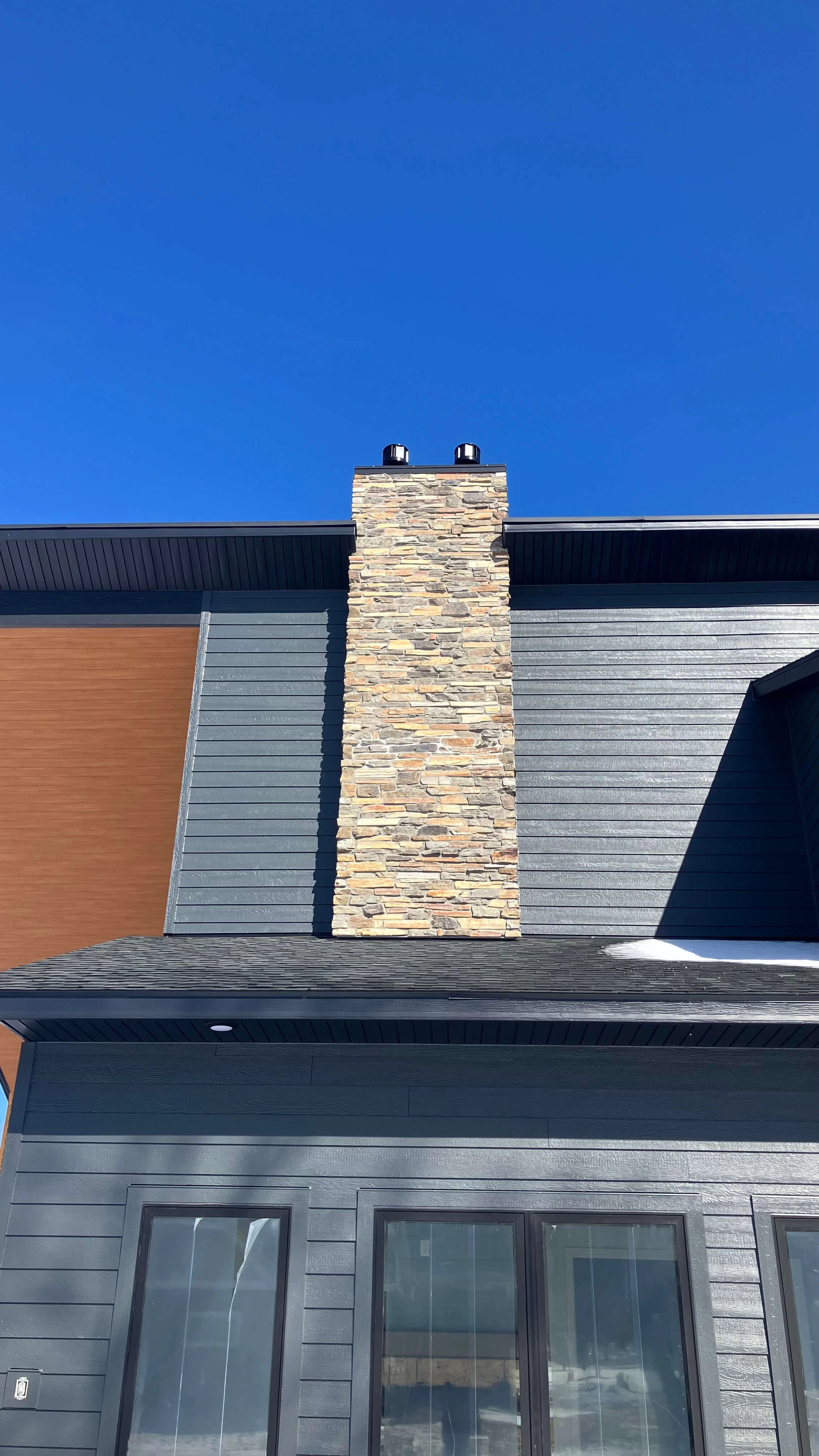 Close-up view of a house with dark gray horizontal siding, a wood accent section, and a stone chimney against a clear blue sky.