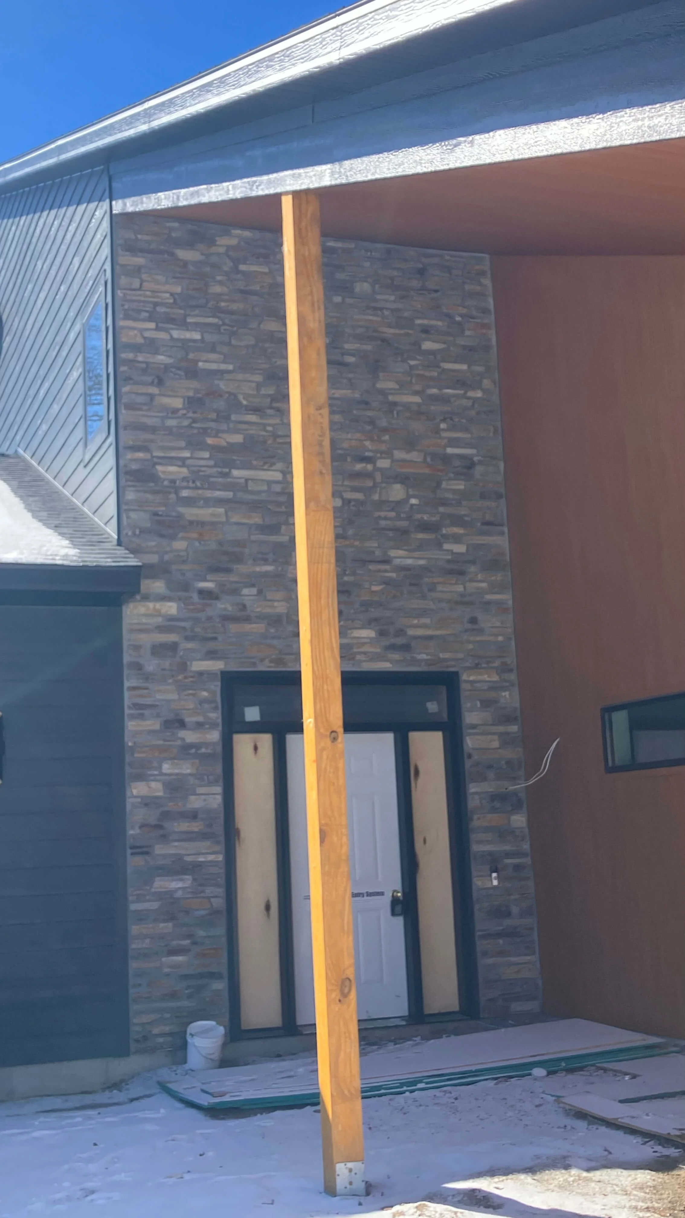 The front of a house under construction, featuring a stone facade, a white door, and a partially built supporting pole in front of the entrance.