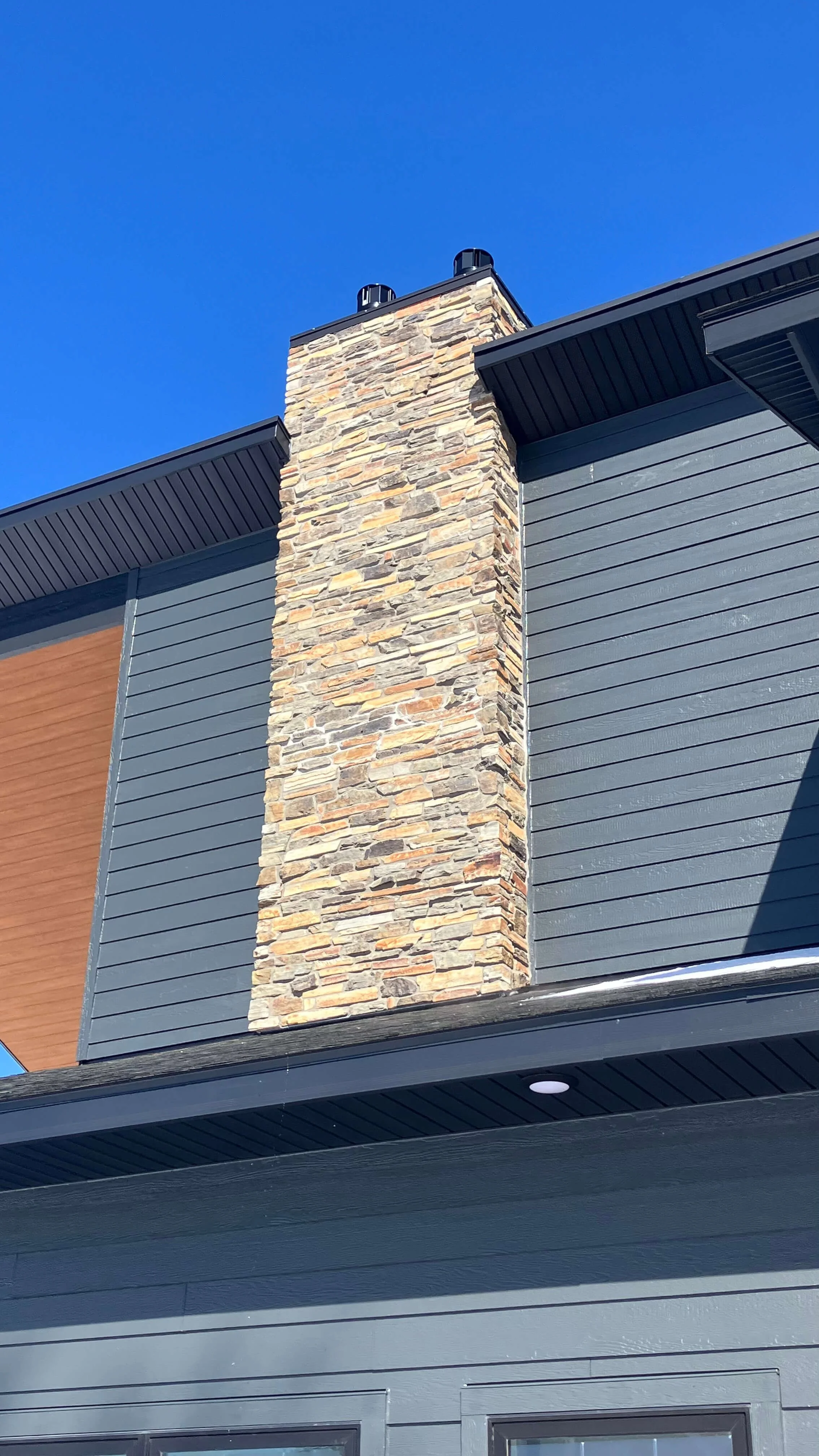 Close-up of a modern house's chimney made of stacked stone, with blue siding and wooden accent panel, under a clear blue sky.