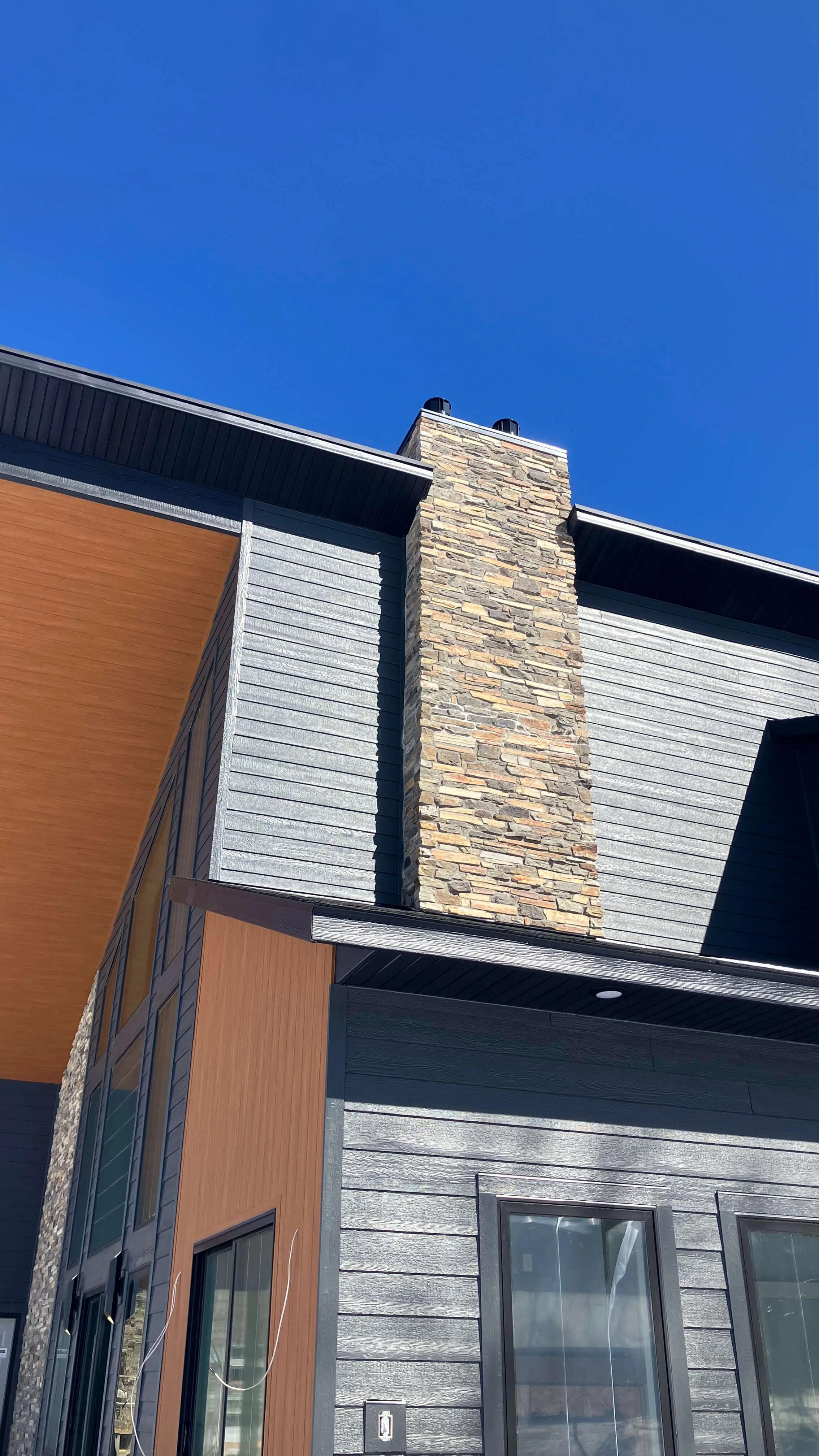 Close-up of a modern house exterior showing a stone chimney, gray siding, and wooden accents against a clear blue sky.