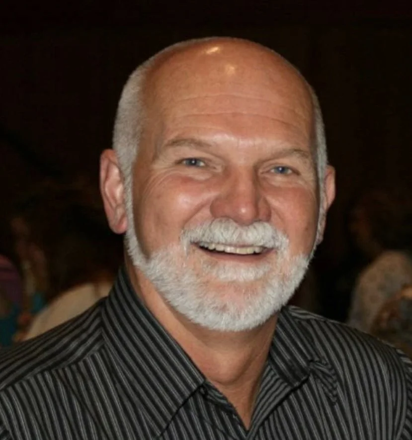 A smiling man with a white beard and bald head, wearing a black and gray striped shirt, at an indoor event.