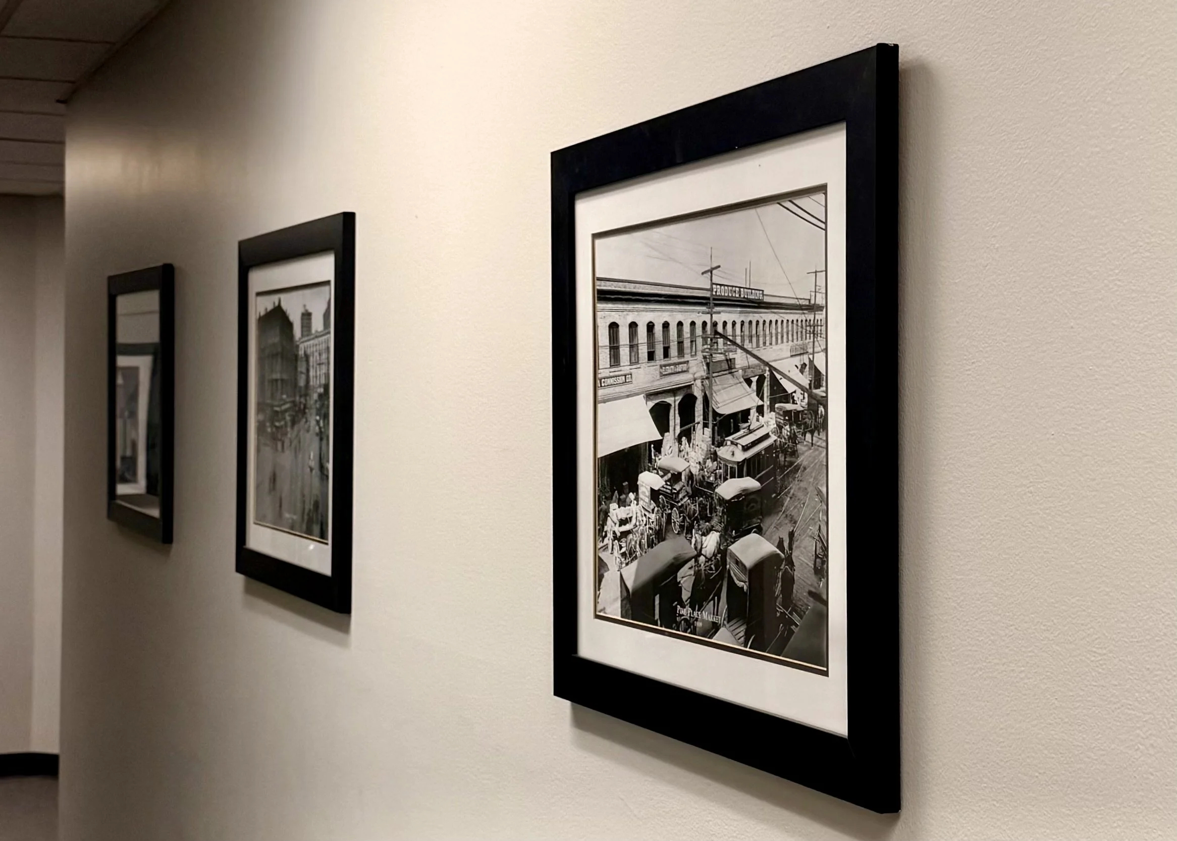 Black-and-white vintage photograph of a busy street scene with horse-drawn carriages and buildings, framed and hanging on a white gallery wall.