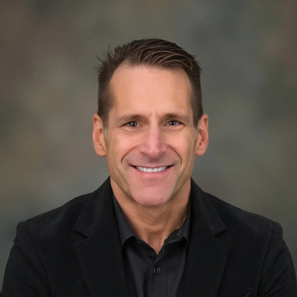 Paul J Casale in a black suit smiling against a blurred gray background.