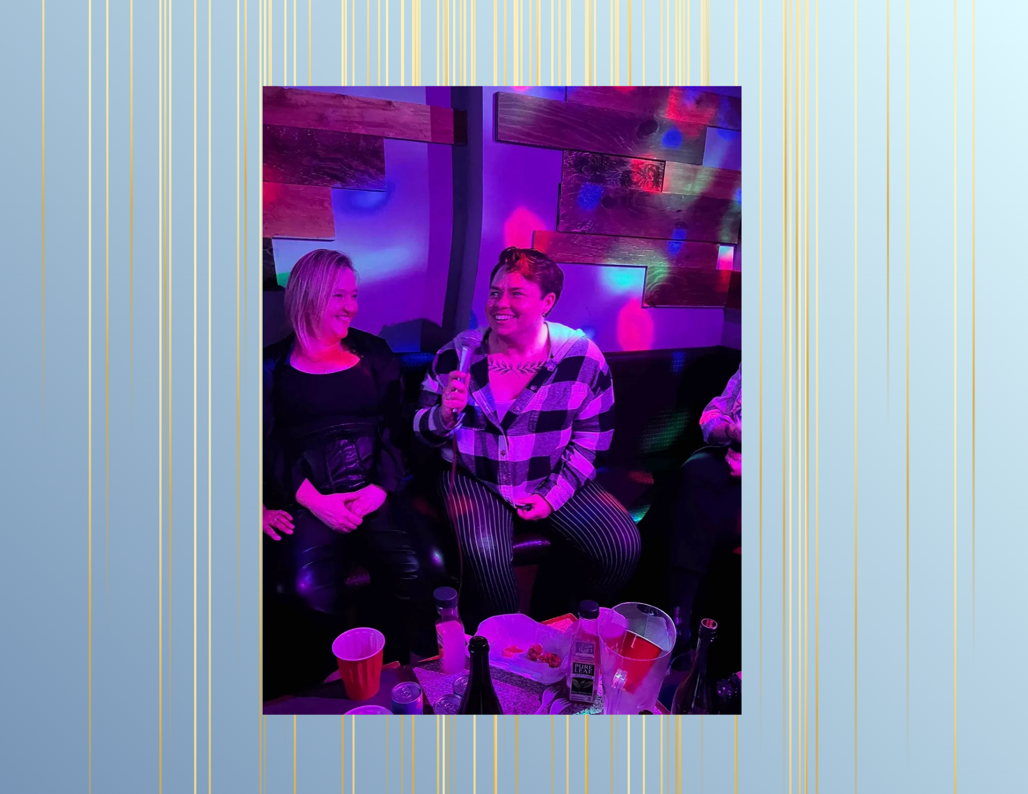 Two women are sitting on a black leather couch in a dimly lit room with colorful purple, pink, and blue lighting. One woman is holding a microphone and smiling, while the other woman is also smiling. There are several bottles, cups, and a bucket on a