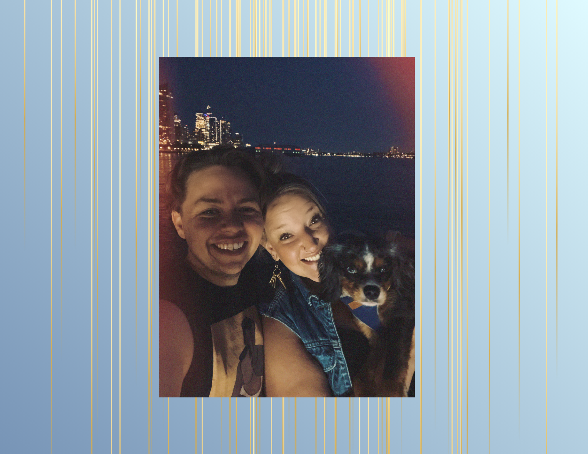 Two smiling women and a dog taking a selfie at night with cityscape and water in the background.
