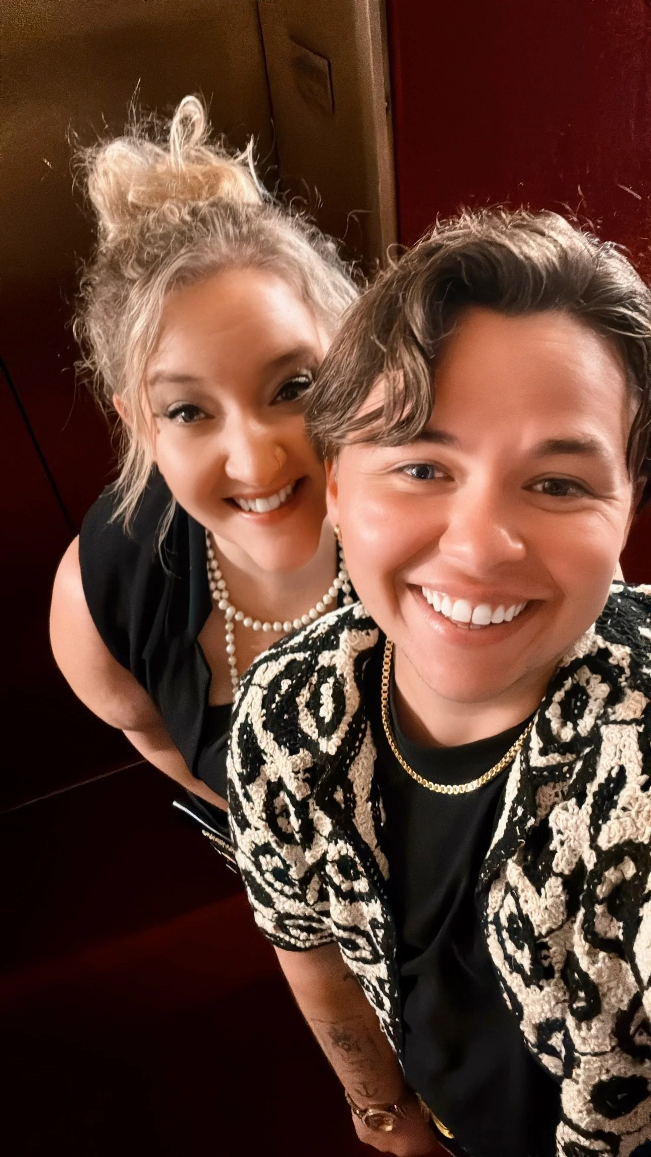 Two women smiling and taking a selfie together, one wearing a black dress with a pearl necklace and the other wearing a patterned jacket and gold chain.