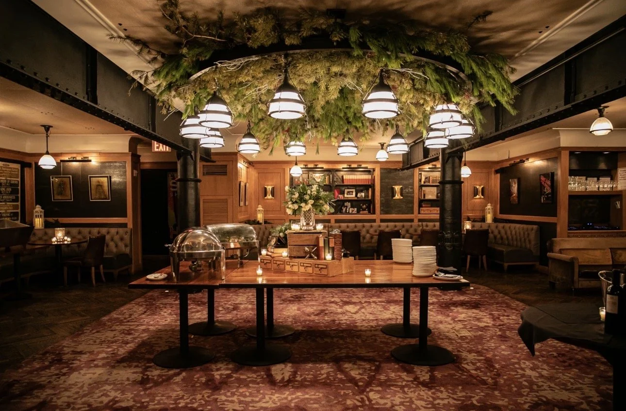 A warmly lit restaurant or pub interior featuring a large wooden table in the center with chafing dishes, surrounded by cozy seating, candles, bookshelves, and decorative greenery hanging from the ceiling.