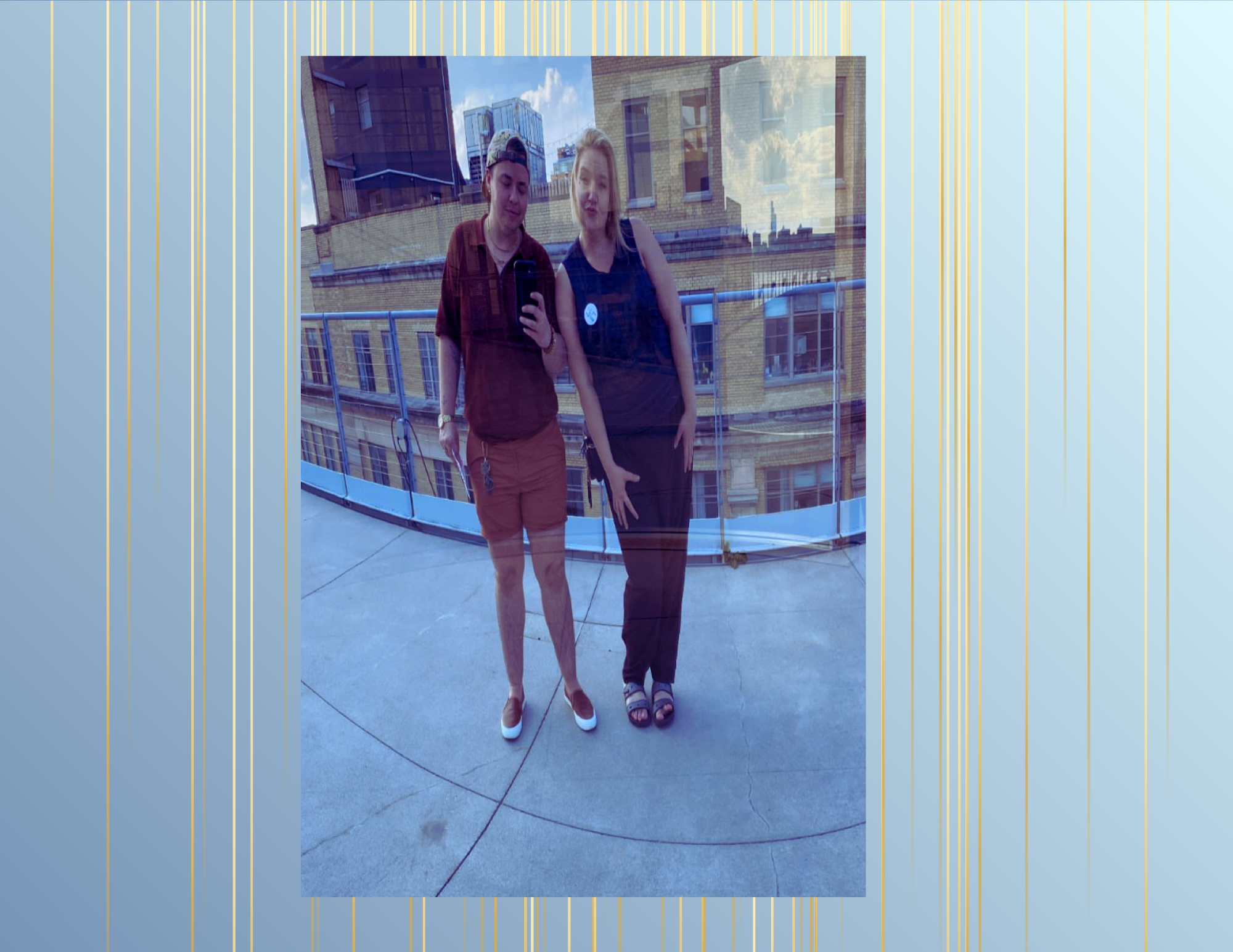 Two people taking a selfie in front of a mirror, with a cityscape reflected in the glass behind them. One is a man in a burgundy shirt and shorts, and the other is a woman in a black outfit.