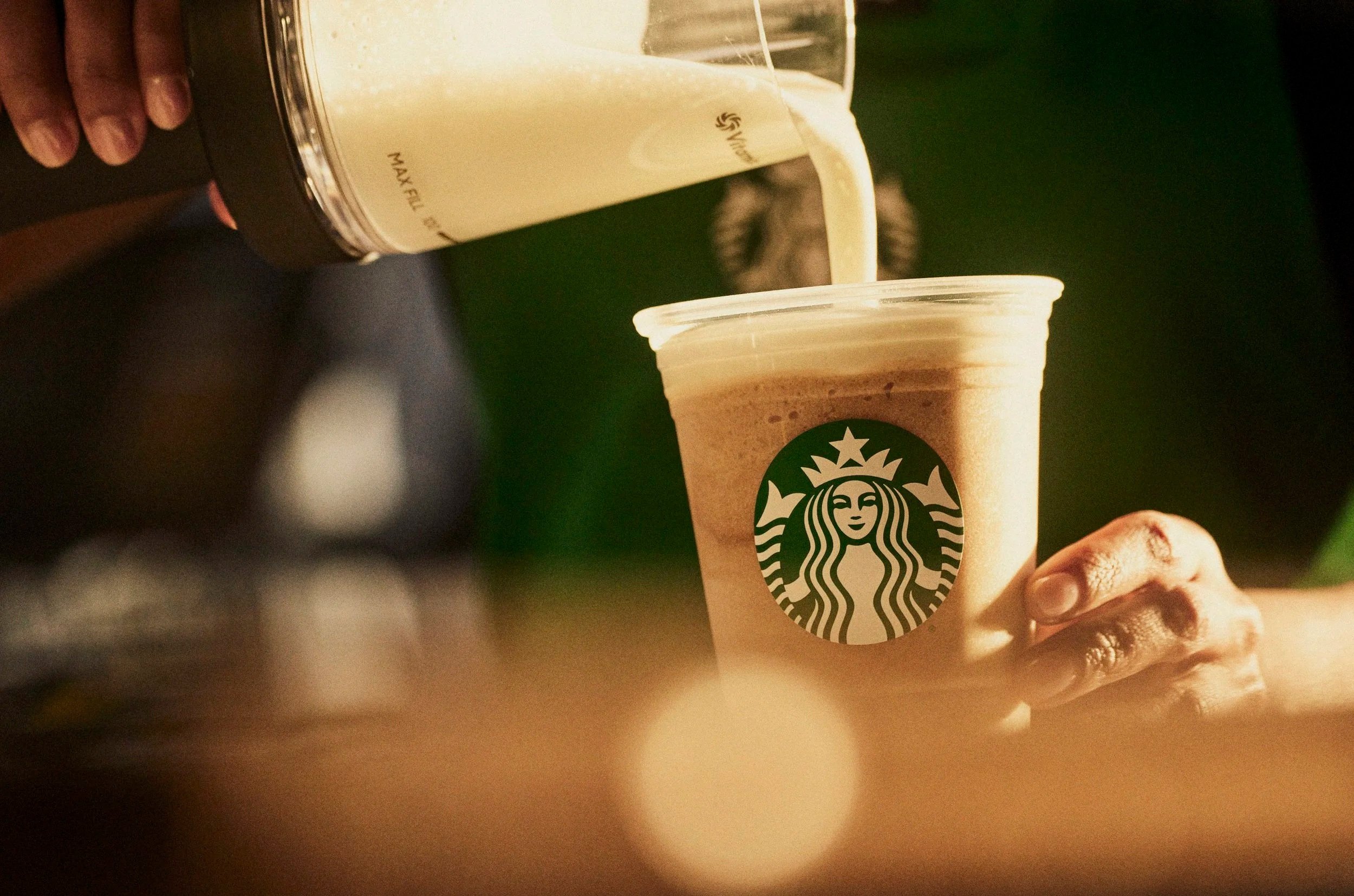 A hand holding a Starbucks cup as coffee is poured from a blender into it, with a green background.