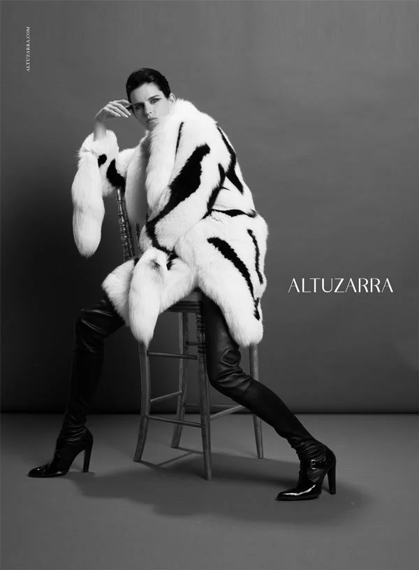 A woman sitting on a wooden chair, wearing a white fur coat with black patterns, black leather pants, and high-heeled boots, posed against a plain background. Altuzzara. Inez and Vinoodh. Stella Tennant.