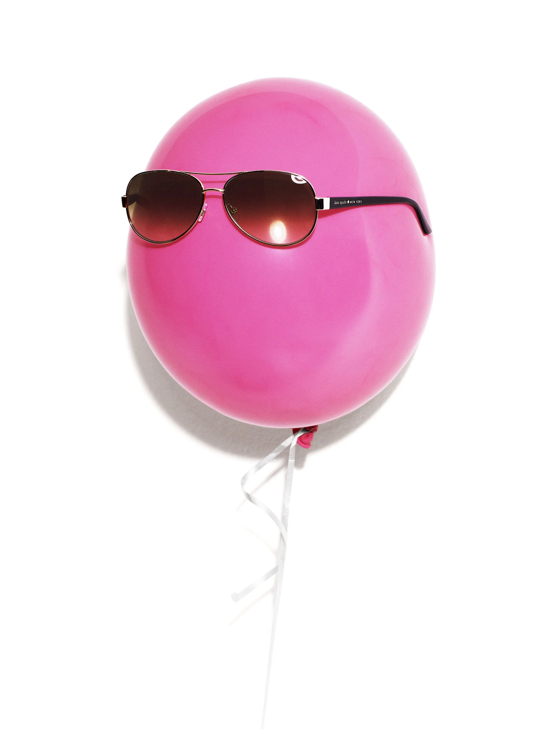 Pink helium balloon wearing a pair of sunglasses with the brand name 'kate spade' on the arm, against a plain white background. Advertising. Producer. Miranda Kendrick. Kate Spade. Bloomingdales.